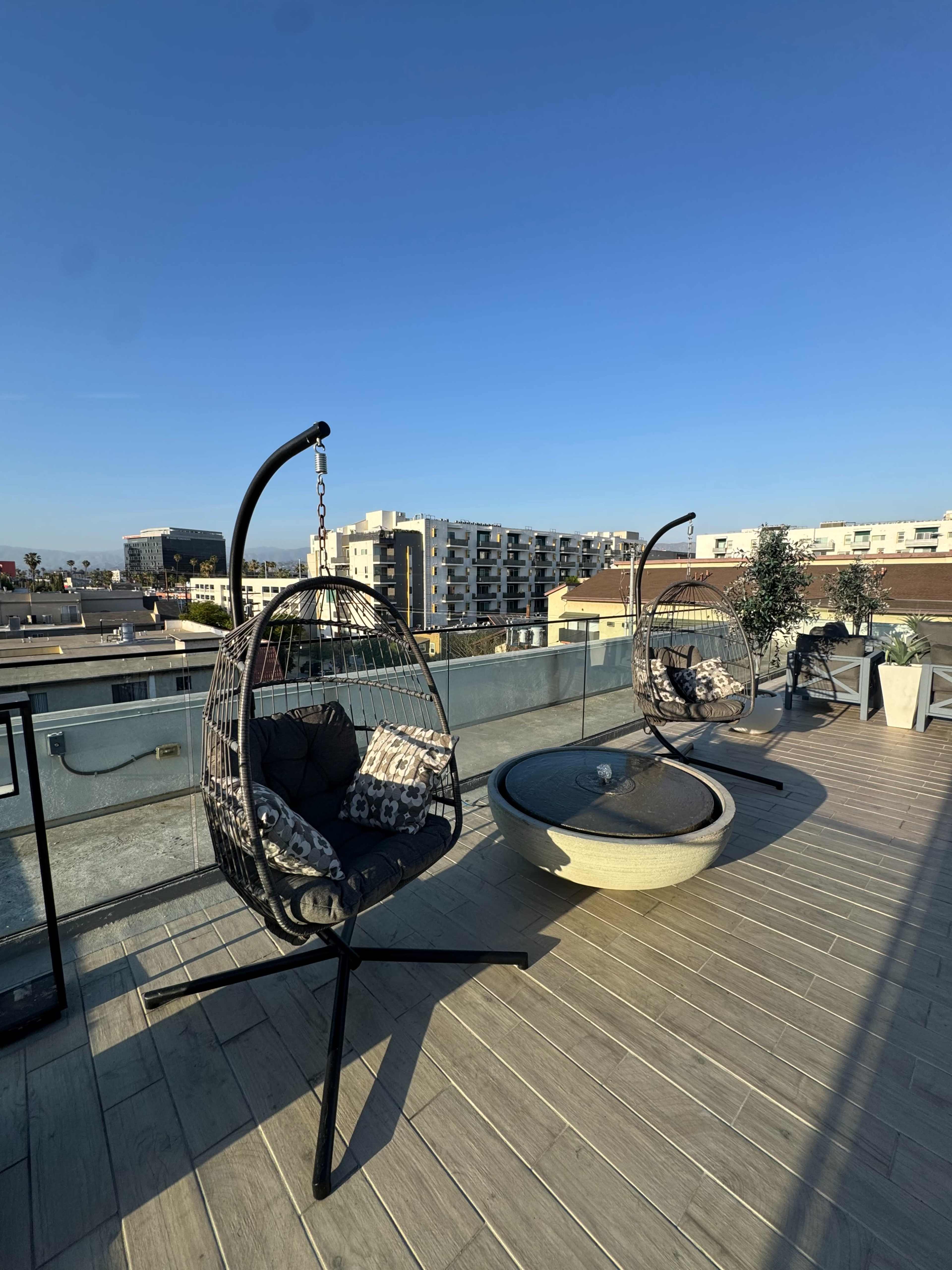 The rooftop features two hanging chairs and a circular fire pit, overlooking a cityscape under a clear blue sky.