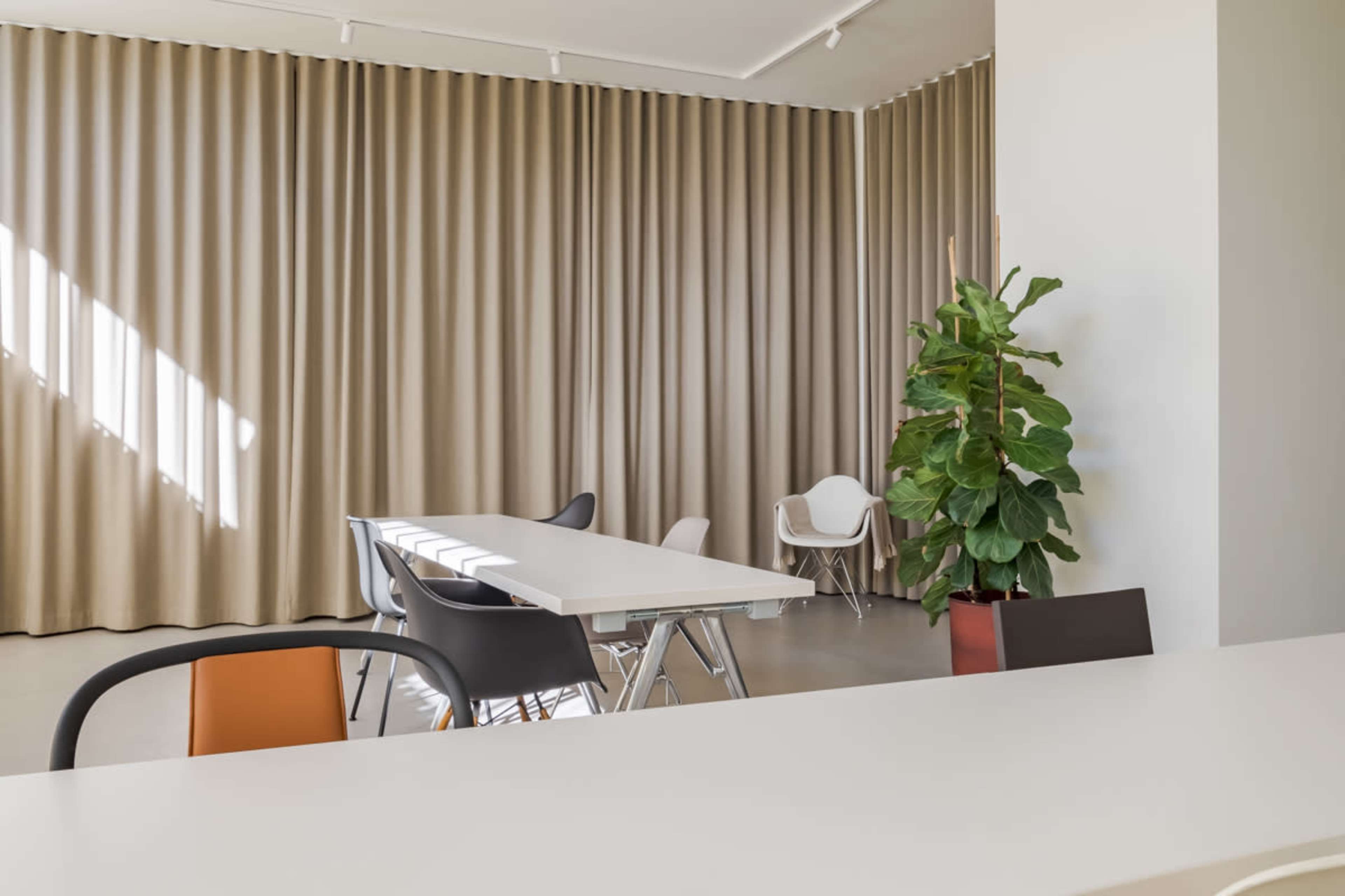A modern room features a white table surrounded by chairs, draped beige curtains, and a potted plant in the corner.