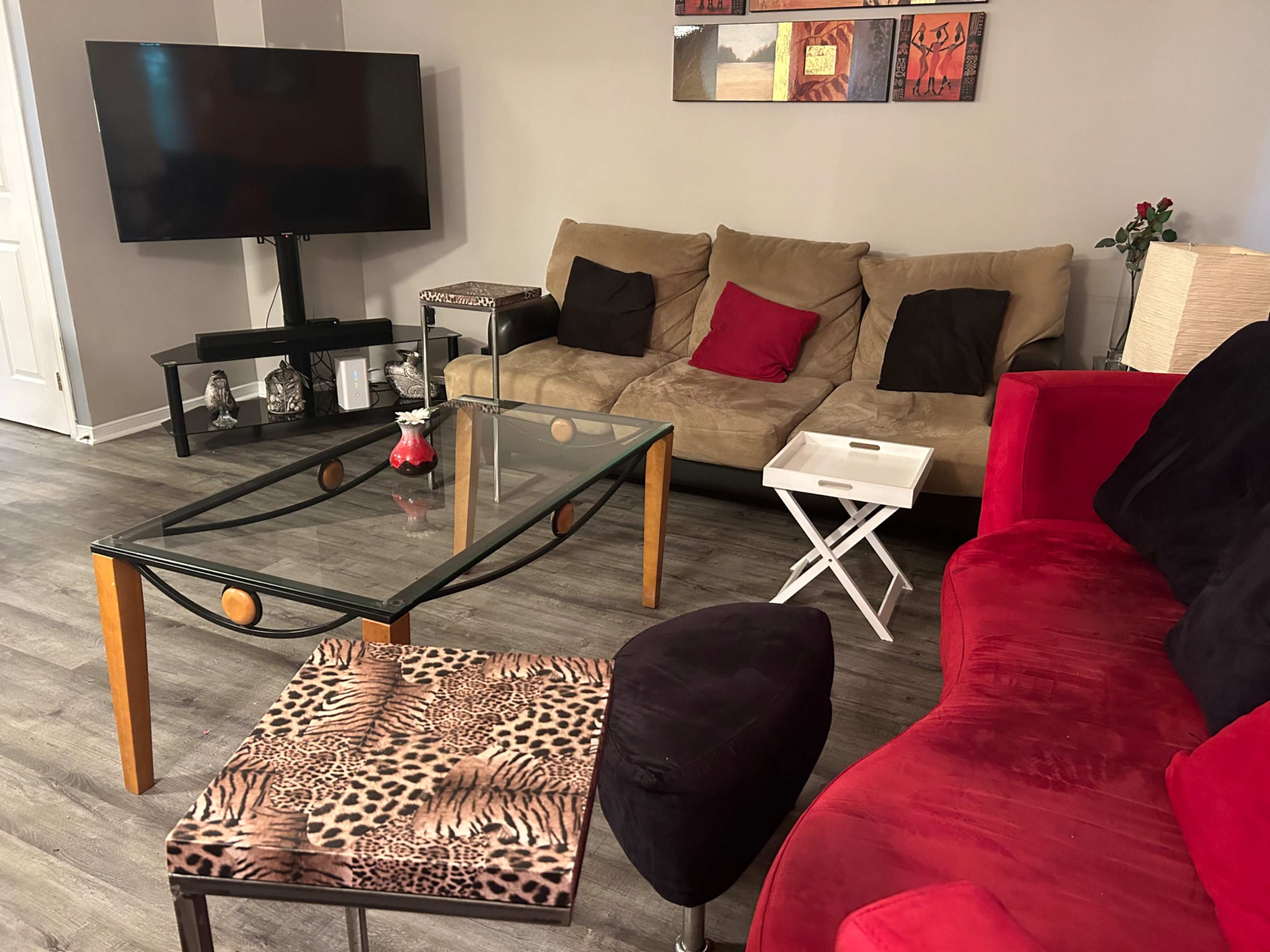 A living room with a beige sofa, a red curved sofa, a glass coffee table with decorative items, a modern TV stand, and a patterned side table.
