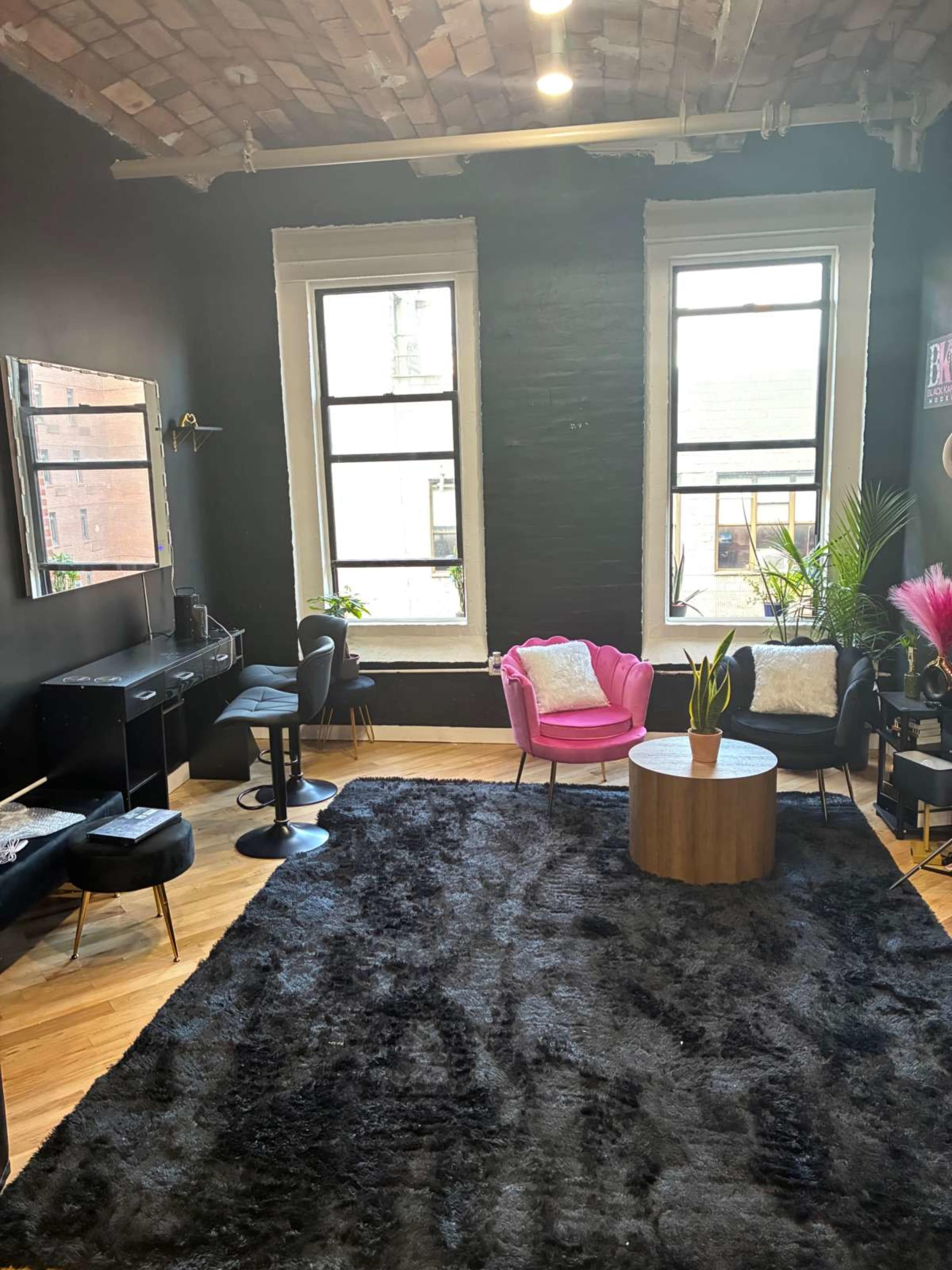 The image shows a modern living room with a large black rug, two black chairs, a pink accent chair, a wooden coffee table, and windows letting in natural light.