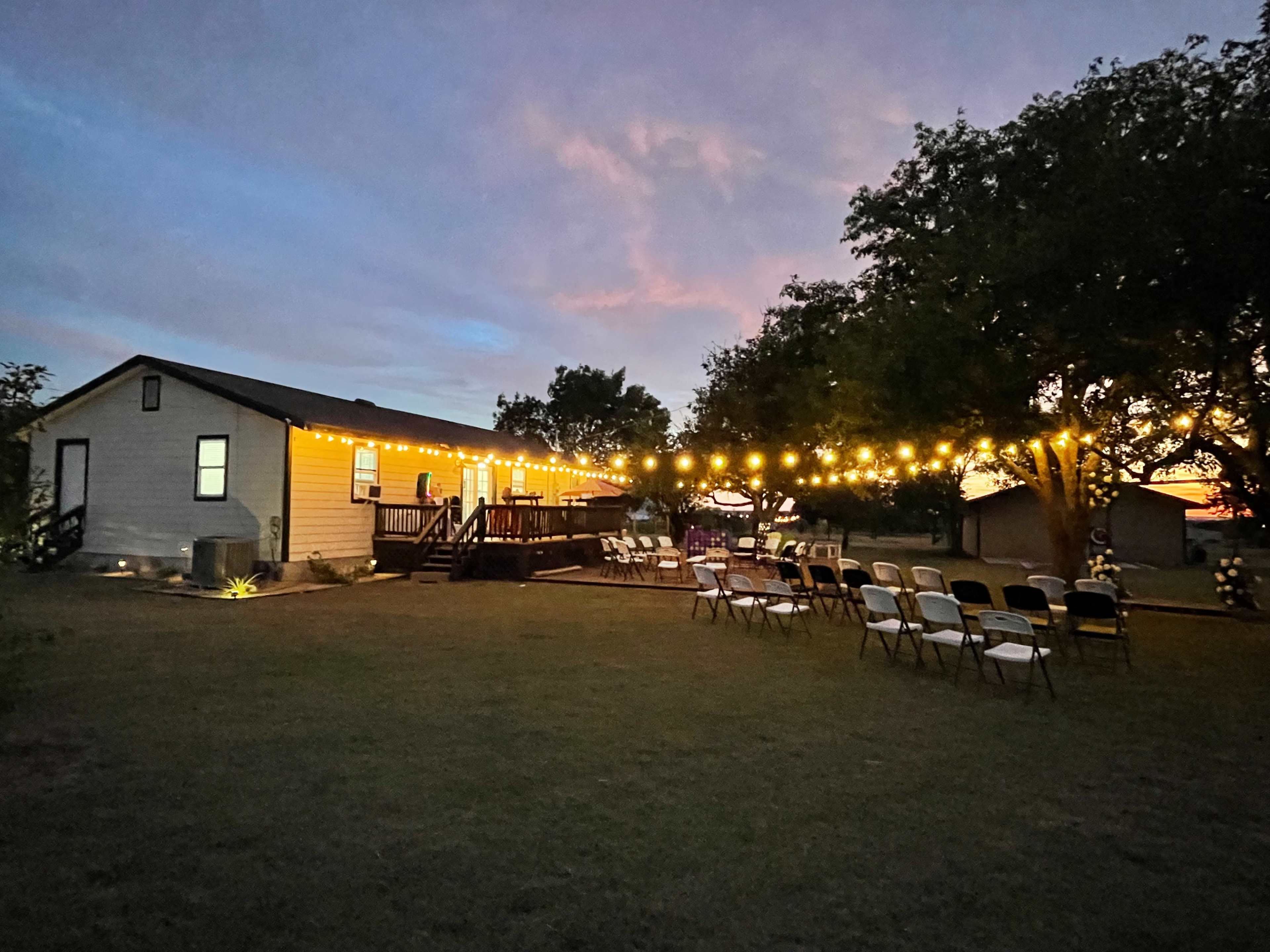 A house with outdoor lighting and rows of chairs set up in a grassy area, indicating a gathering space during dusk.