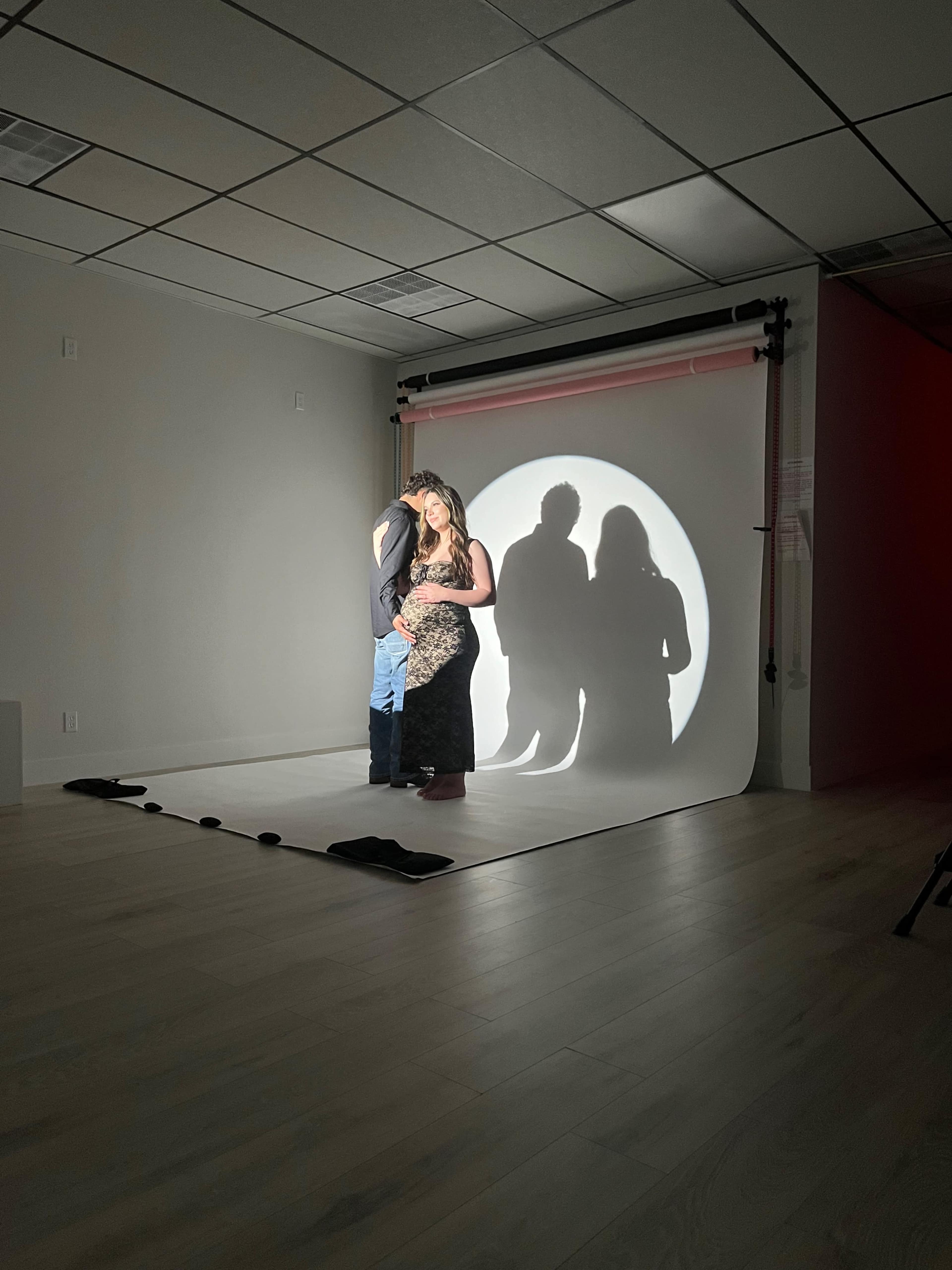A couple stands closely together in front of a circular light projection, casting shadows on a backdrop in a photography studio.