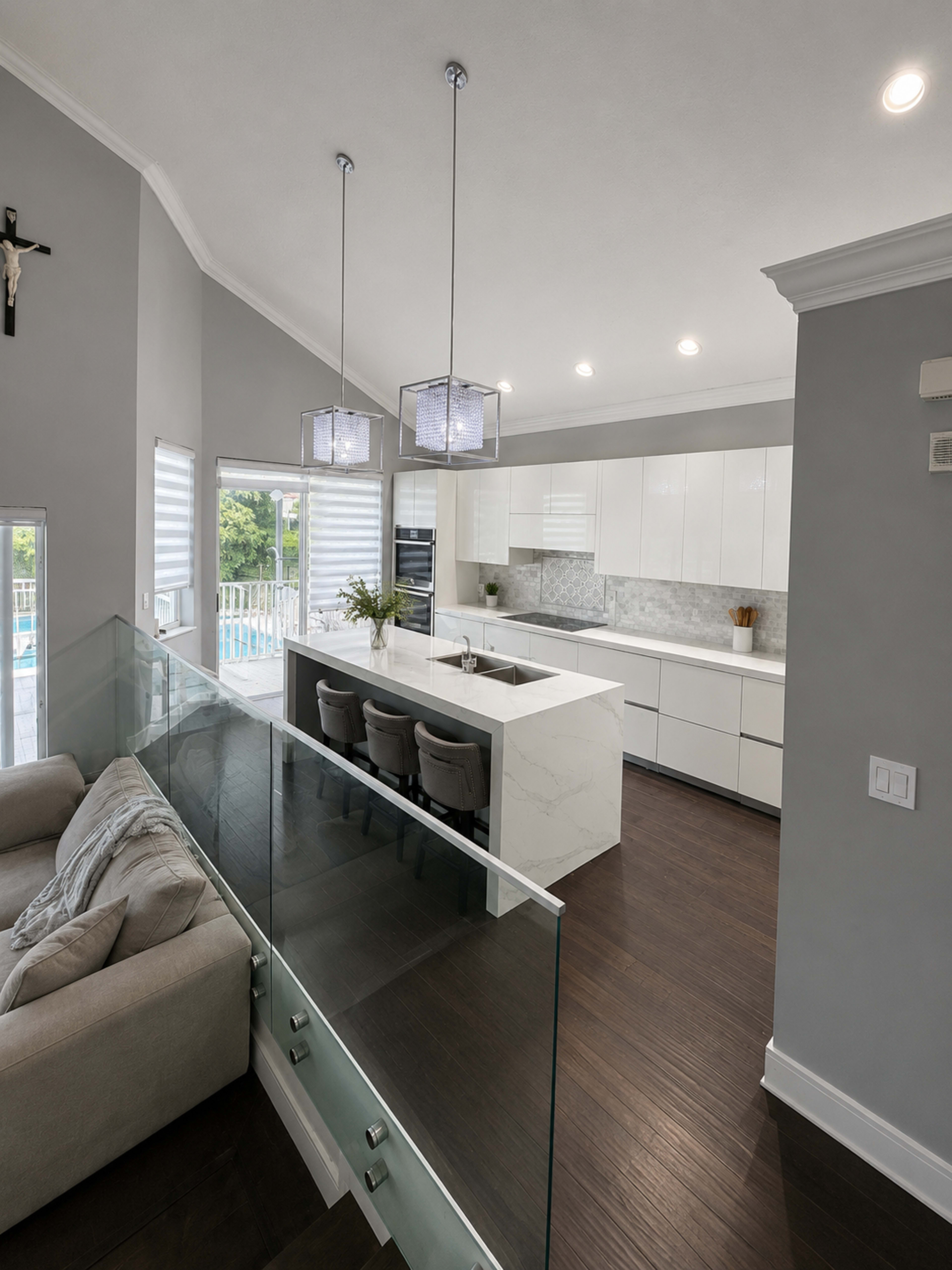 The image shows a modern kitchen with white cabinetry, a large island, and pendant lighting, overlooking a living space with a glass railing.