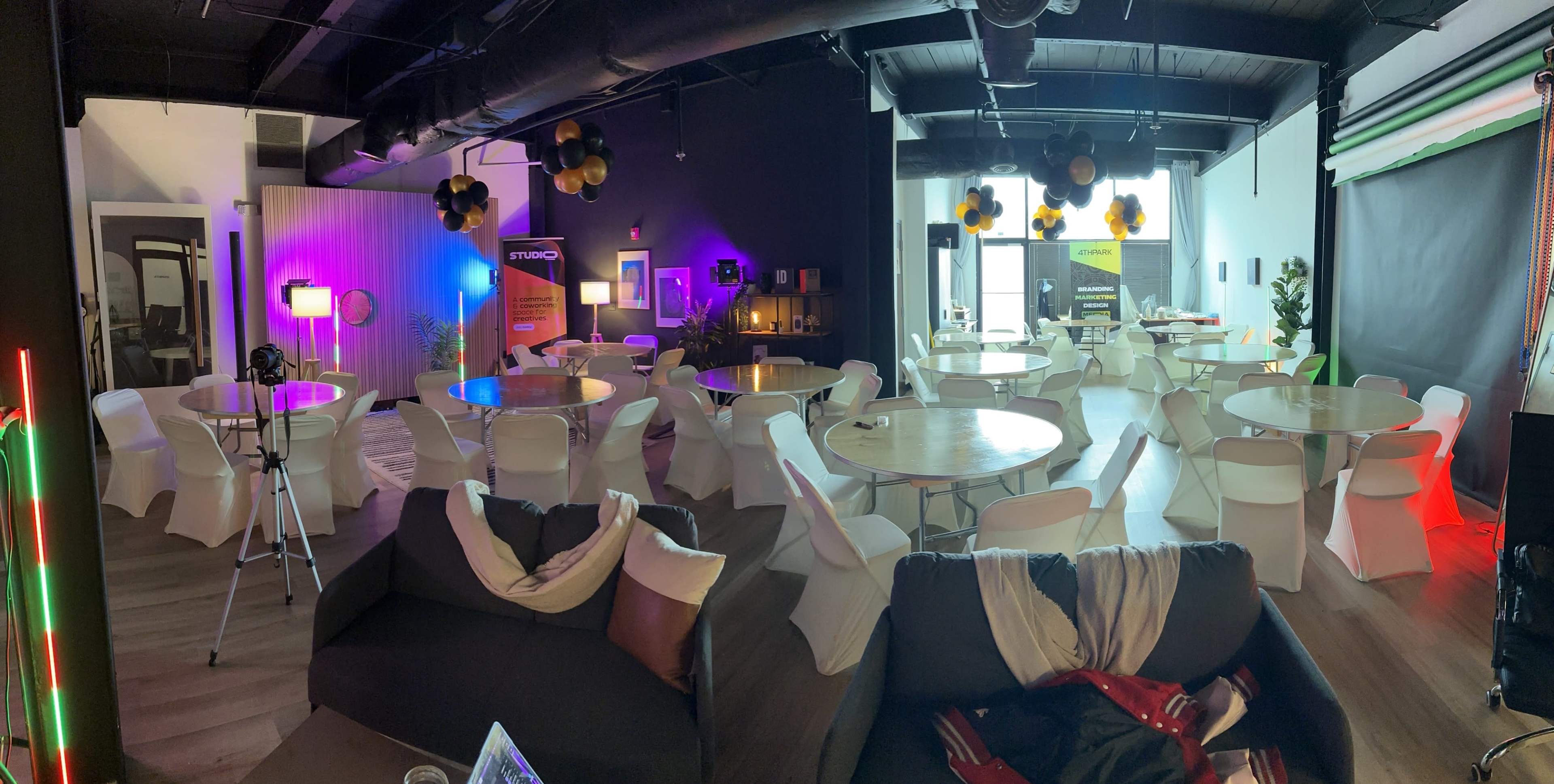 A spacious event area with round tables and white chairs, decorated with black and yellow balloons and warm lighting, suitable for gatherings or presentations.