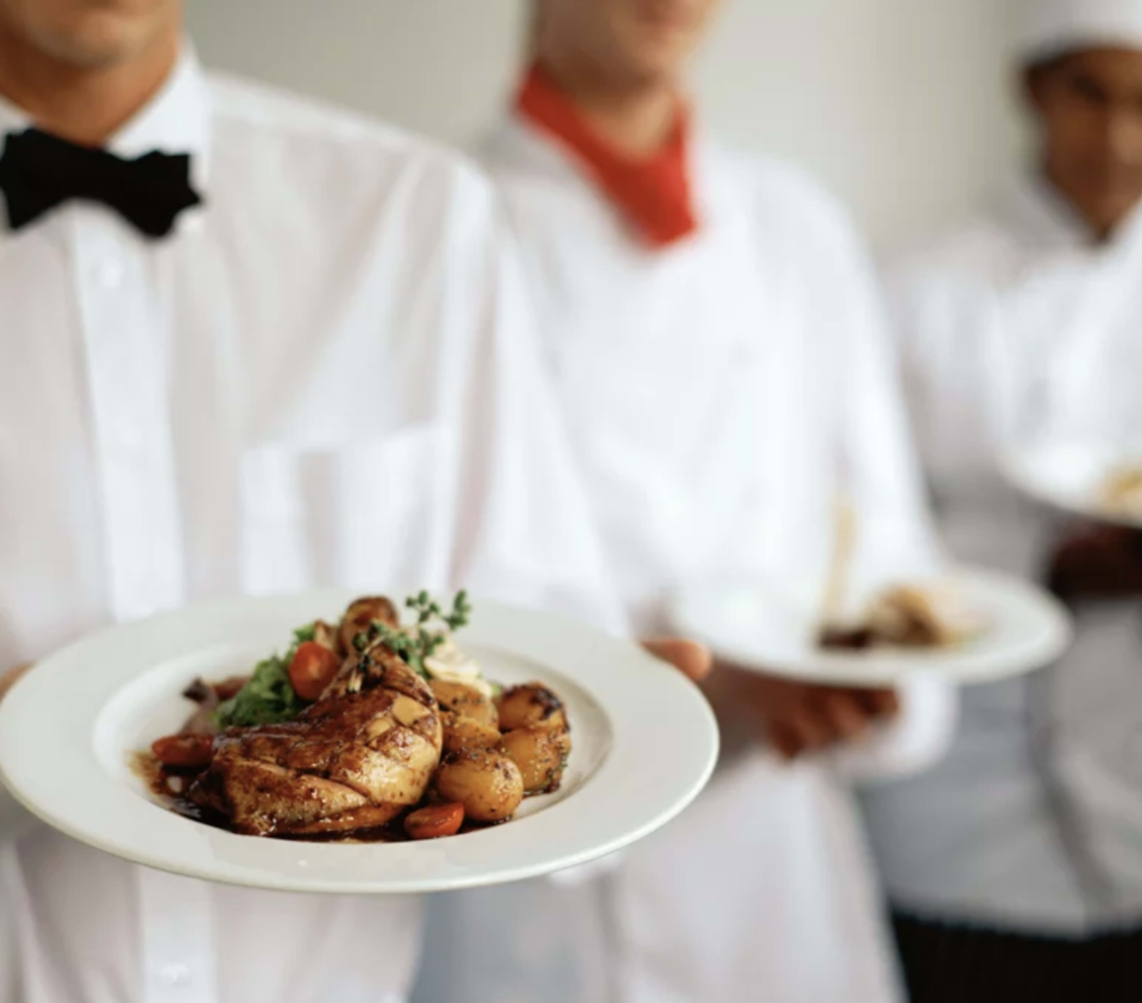 Three chefs in white uniforms hold plates of food, with one chef in the foreground presenting a dish featuring roasted chicken, potatoes, and garnishes.