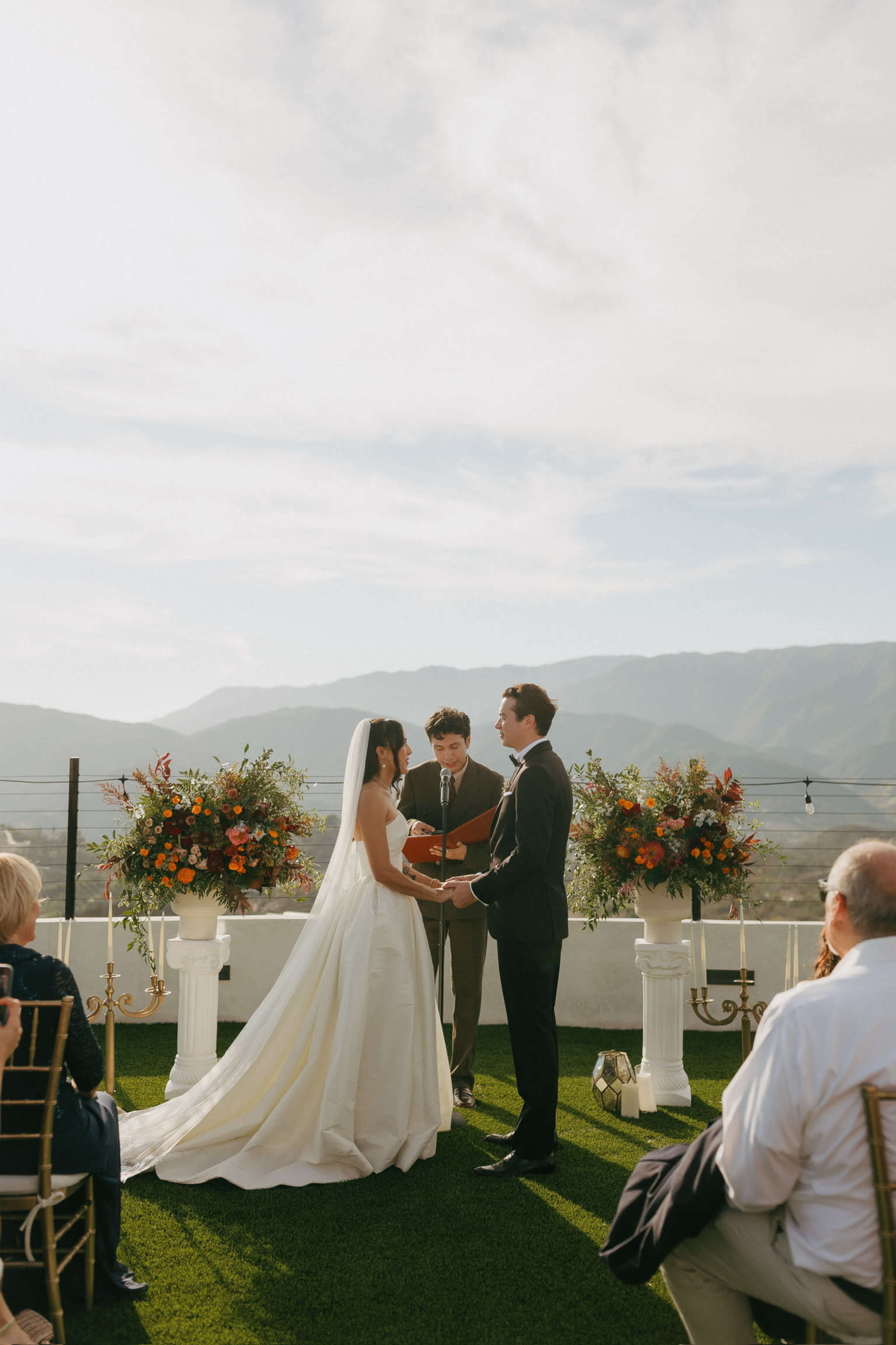 A couple stands in front of an officiant on a terrace adorned with floral arrangements while guests observe in the background.