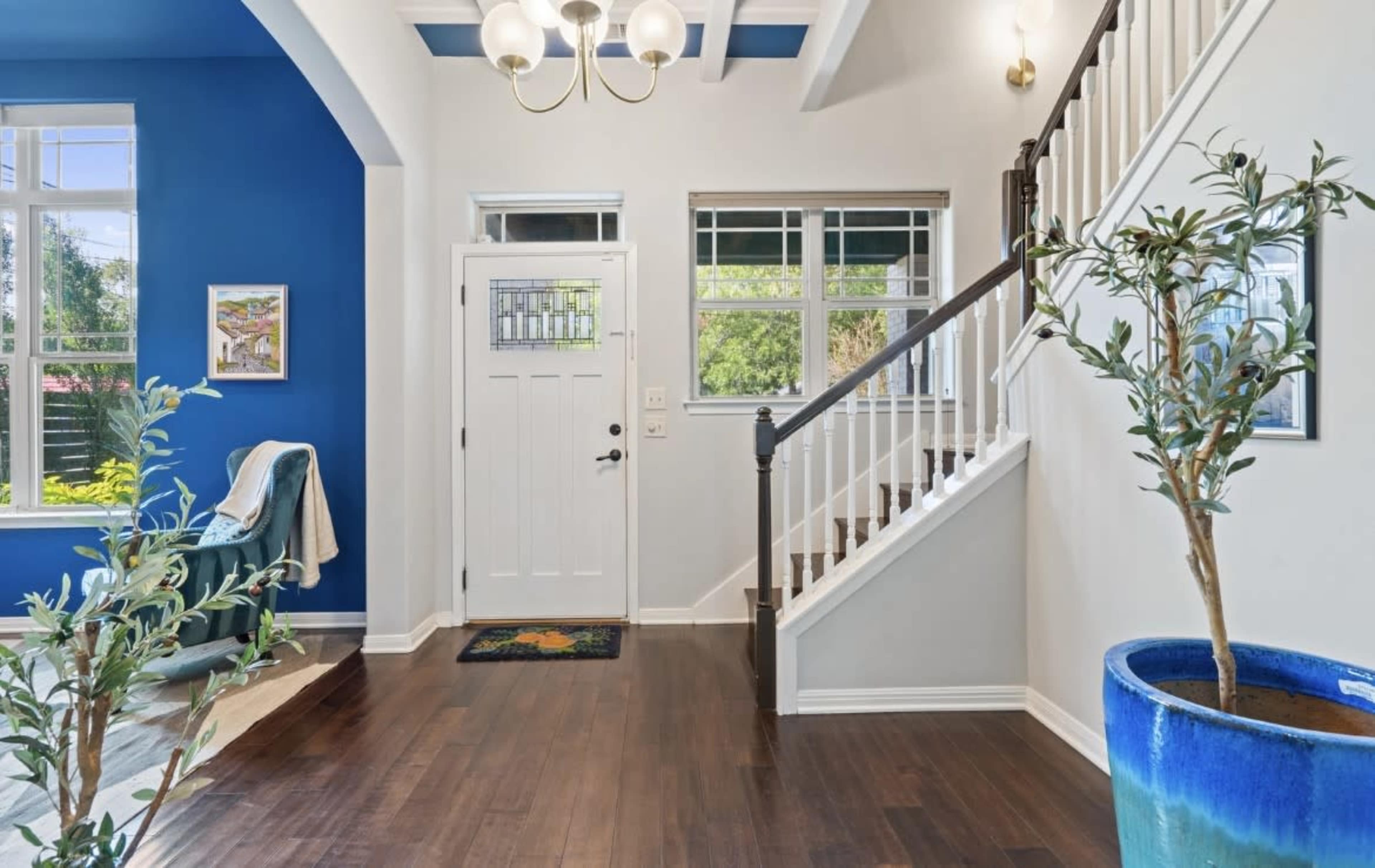 The image shows a well-lit entryway with a staircase, a blue accent wall, a front door, and potted plants.