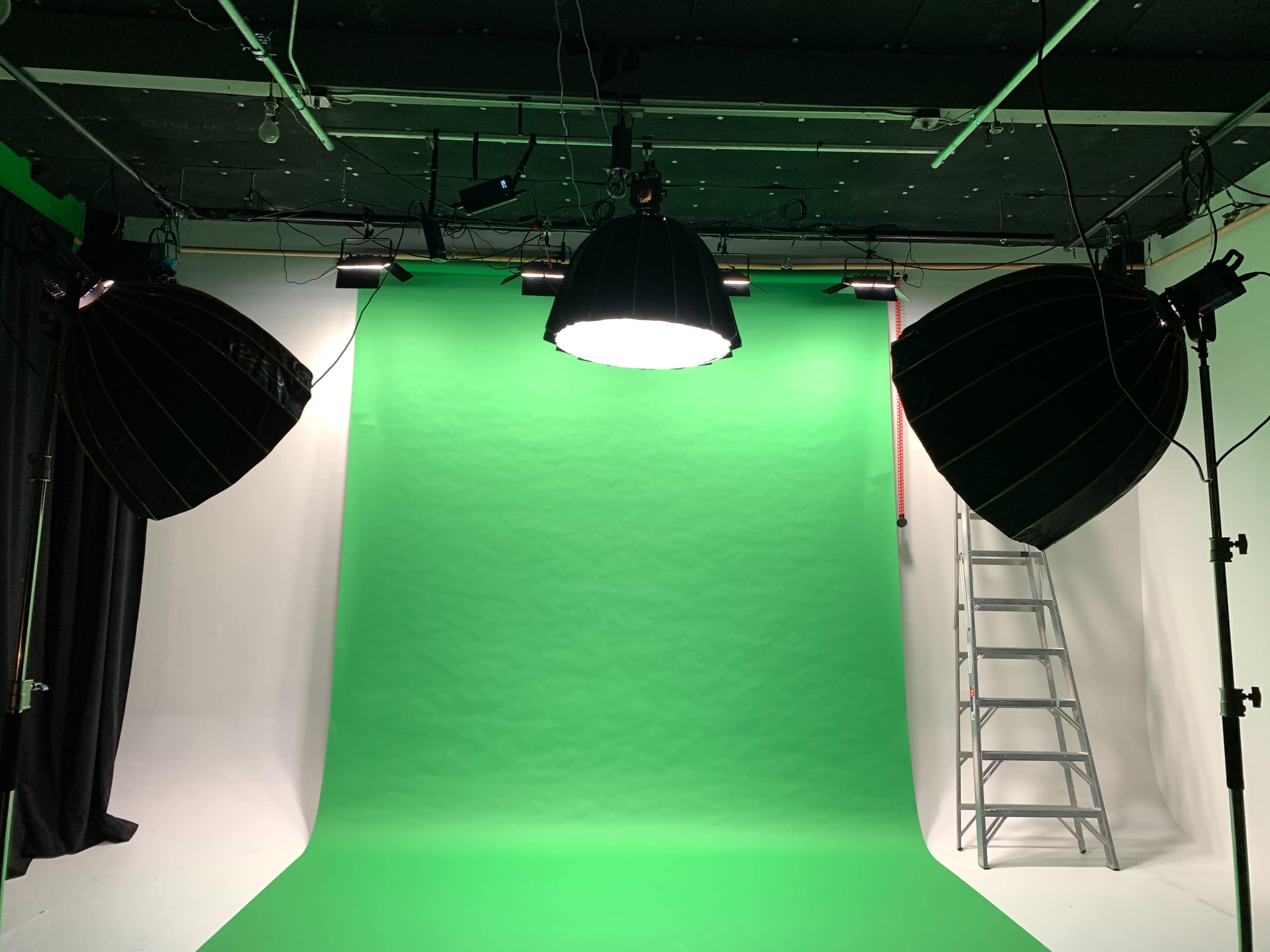 A photography studio with green screens, two large softbox lights, and a ladder in the background.