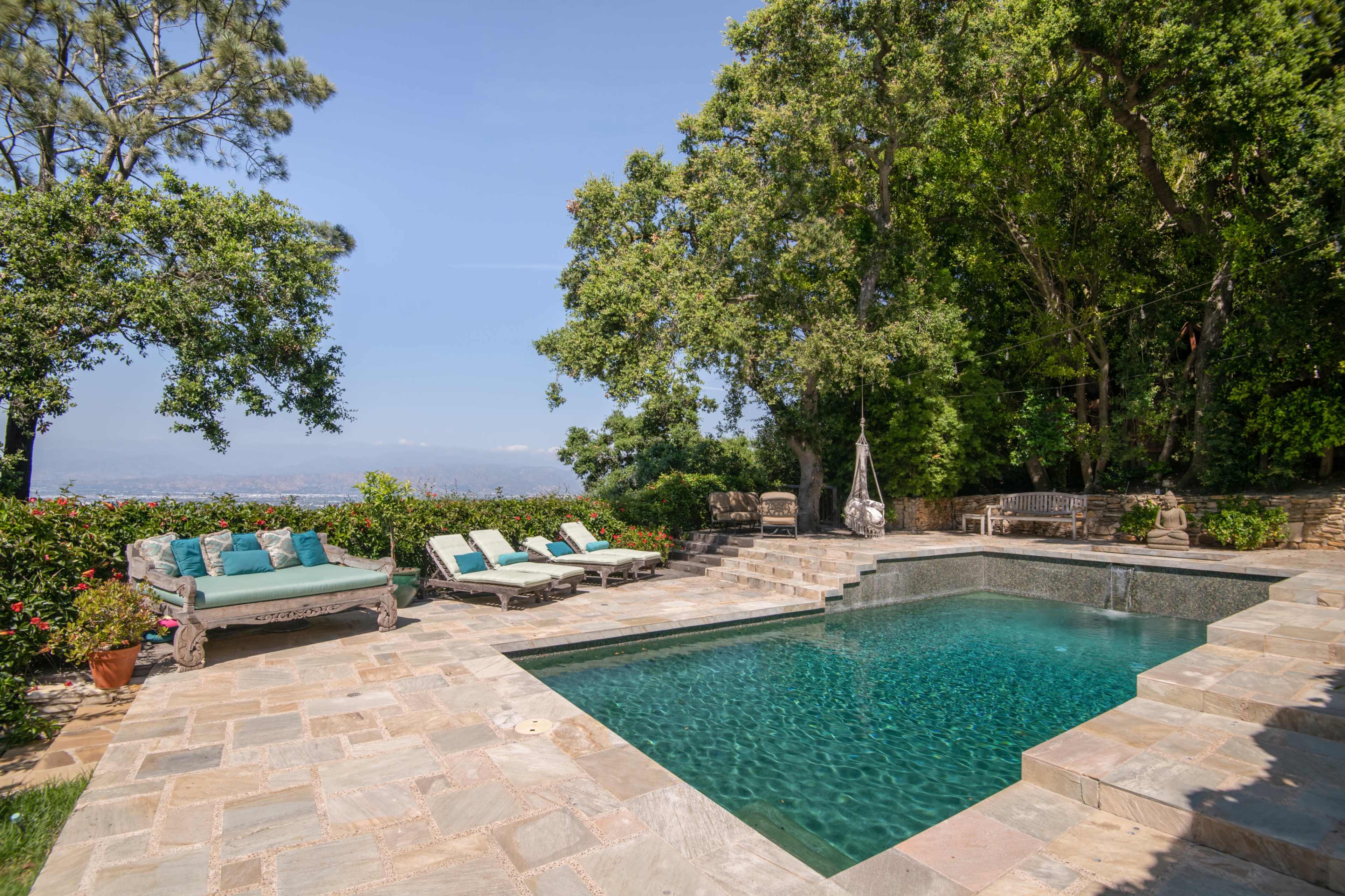 A rectangular swimming pool is bordered by a stone patio and lounge chairs, surrounded by trees and flowering plants.