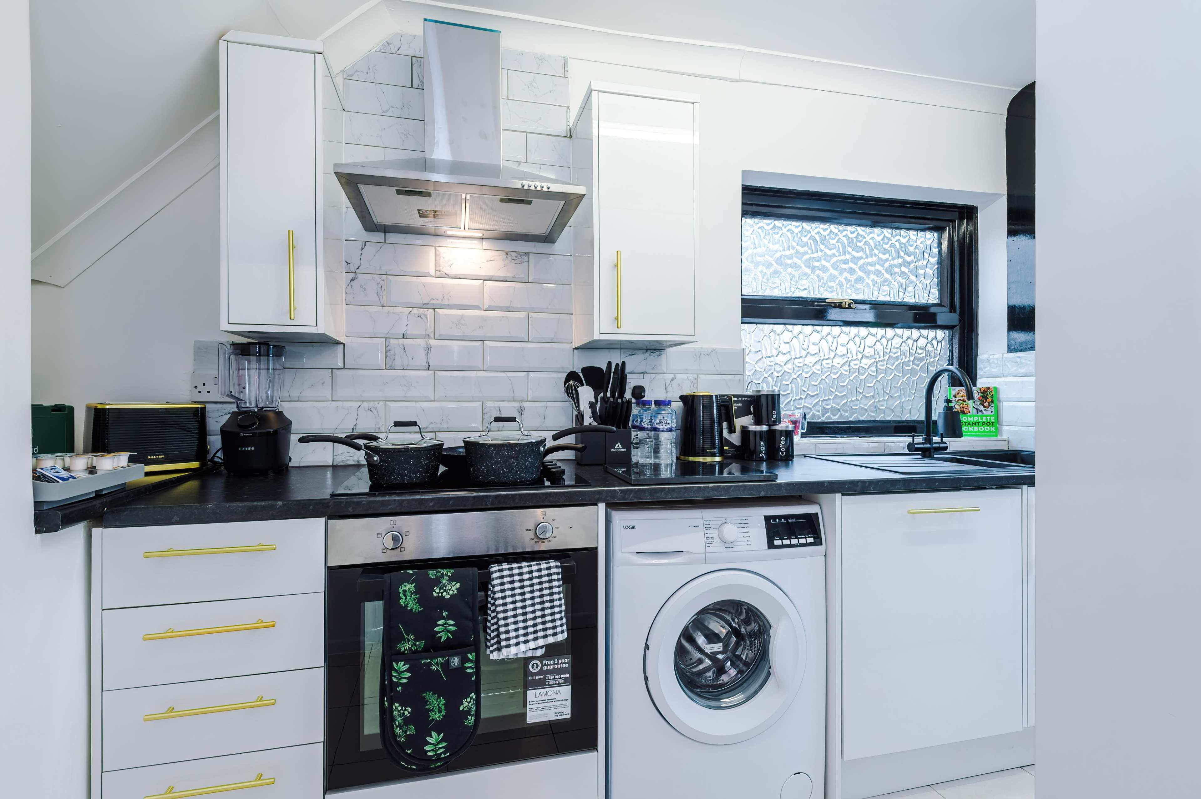 A modern kitchen features white cabinets, a black countertop, two cooking pots on a stove, a washing machine, and a window with textured glass.