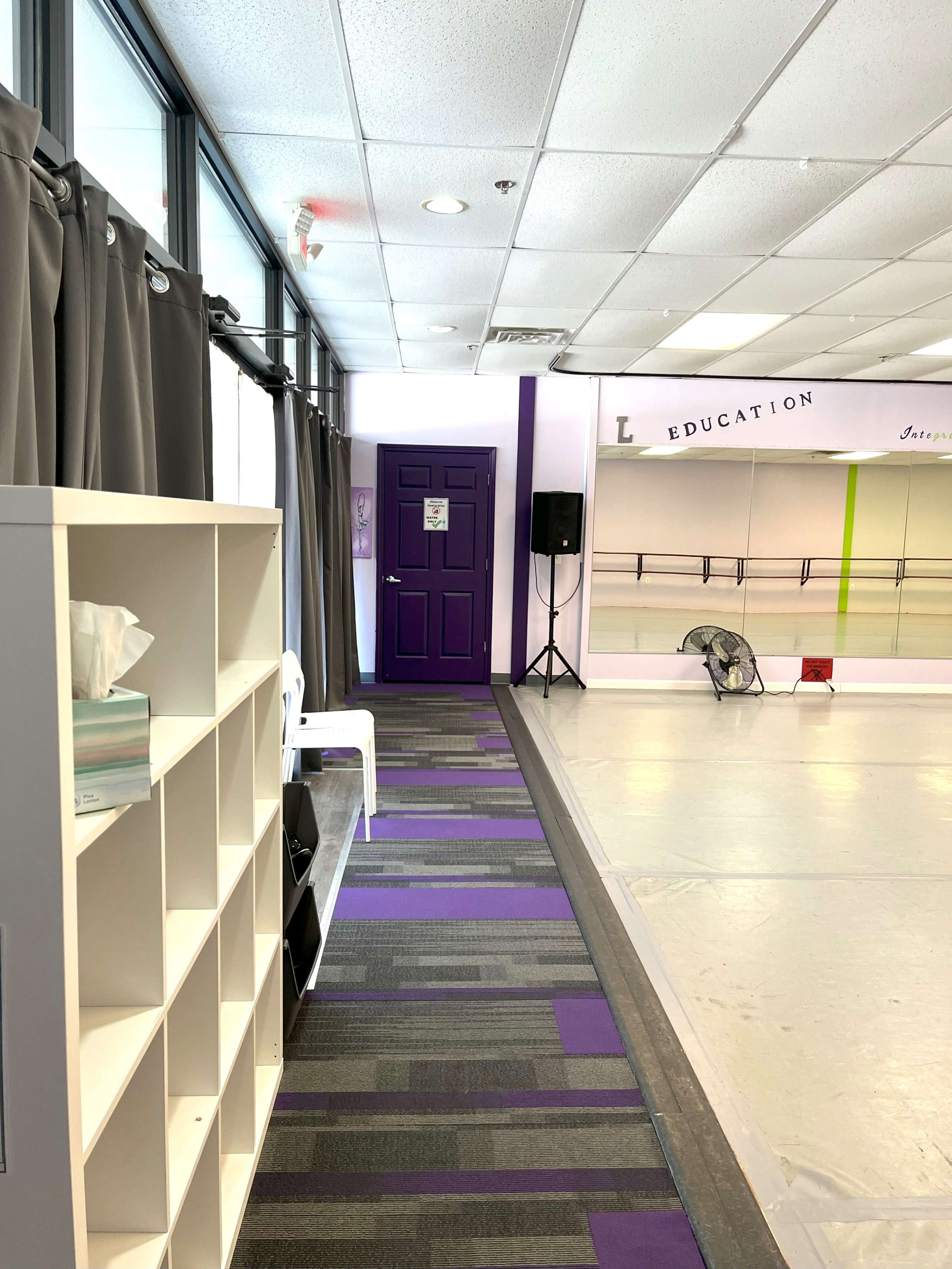 A hallway leads to a dance studio, featuring a purple door, gray curtains, and a white shelving unit along one side.