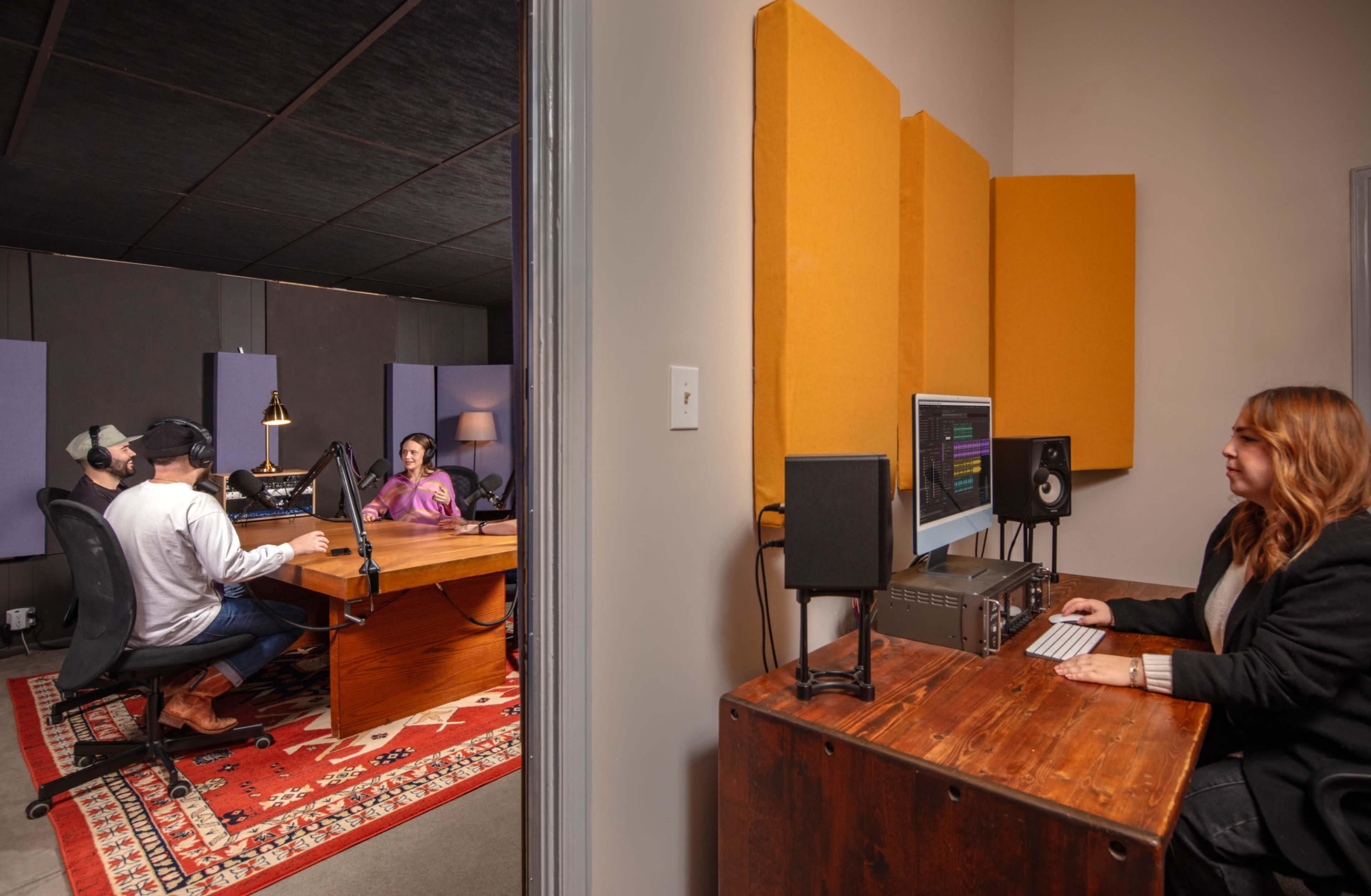 A recording studio is shown with two individuals at a table discussing, while a third person works on a computer in a separate area.