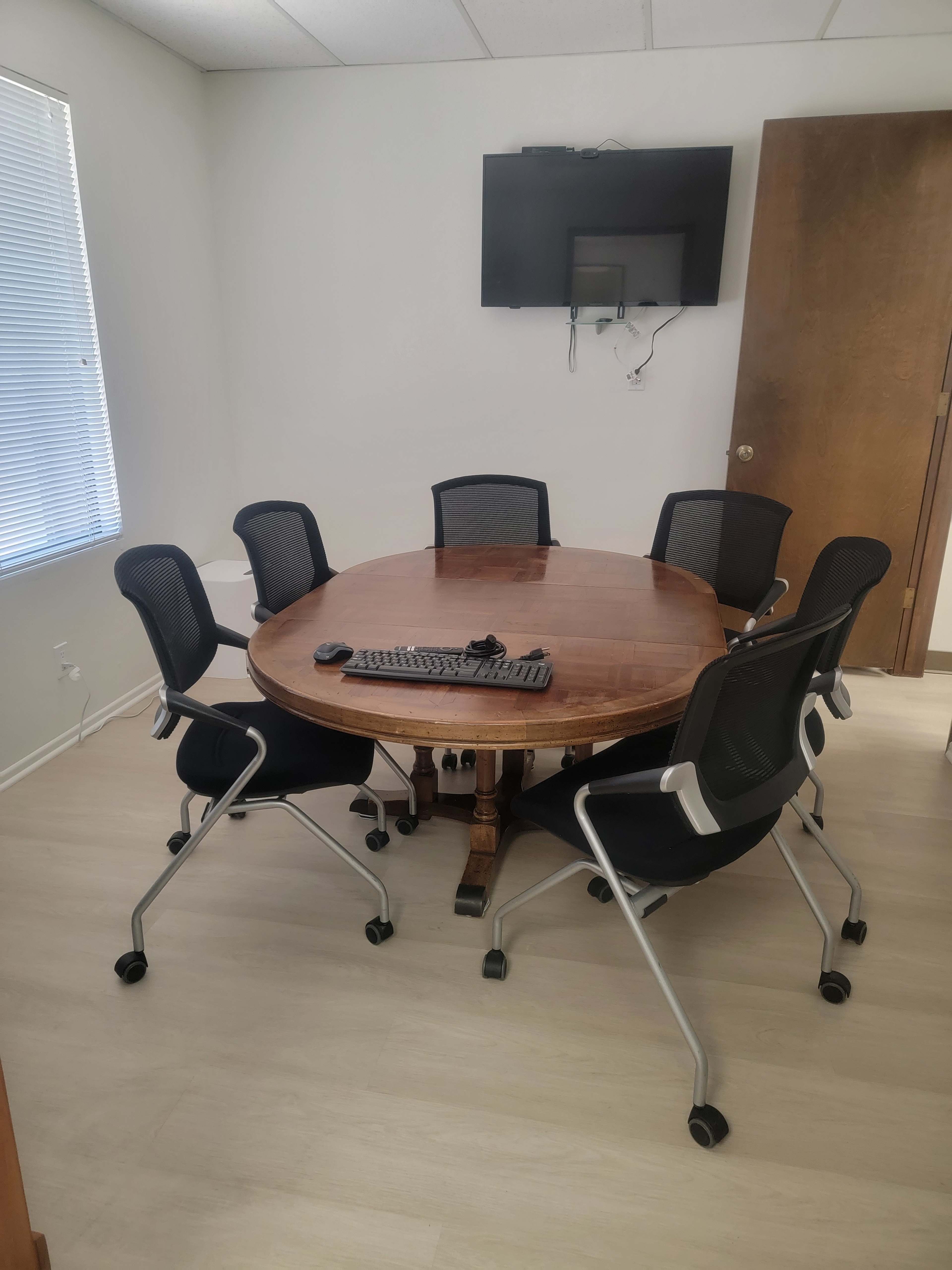 A round wooden table surrounded by six black rolling chairs is positioned in a well-lit meeting room, with a television mounted on the wall.