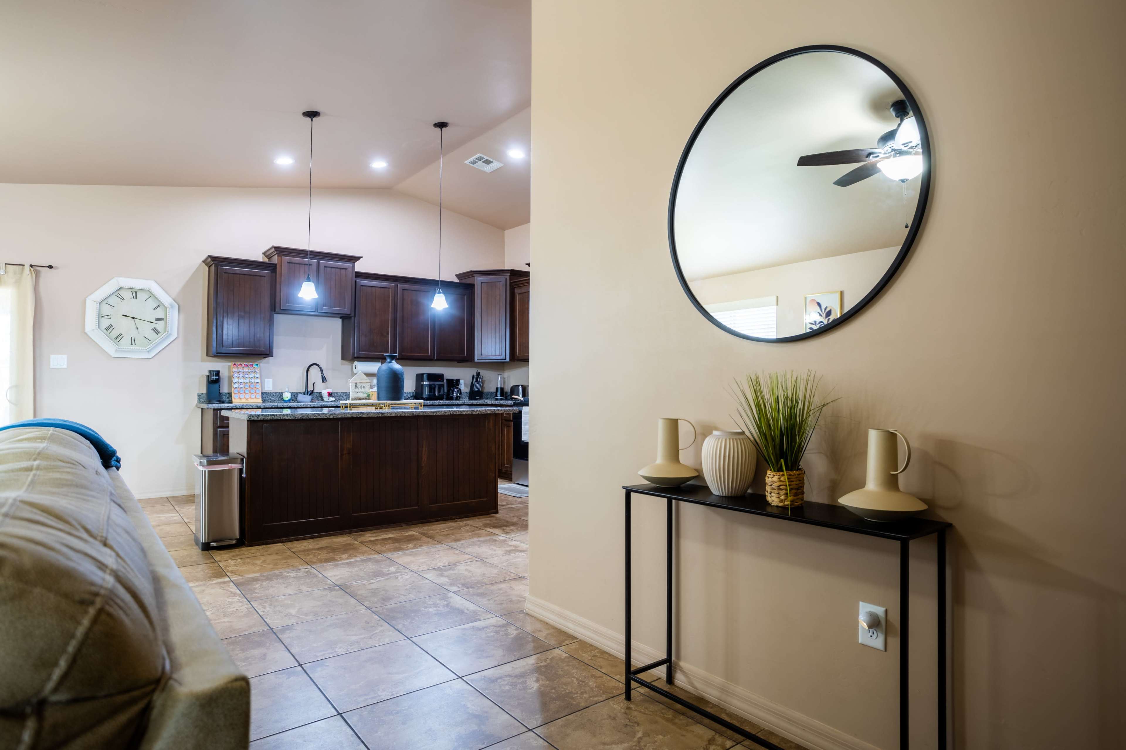 A living area features a beige wall, a round mirror on a console table, and a view of a modern kitchen with dark wood cabinets and pendant lights.