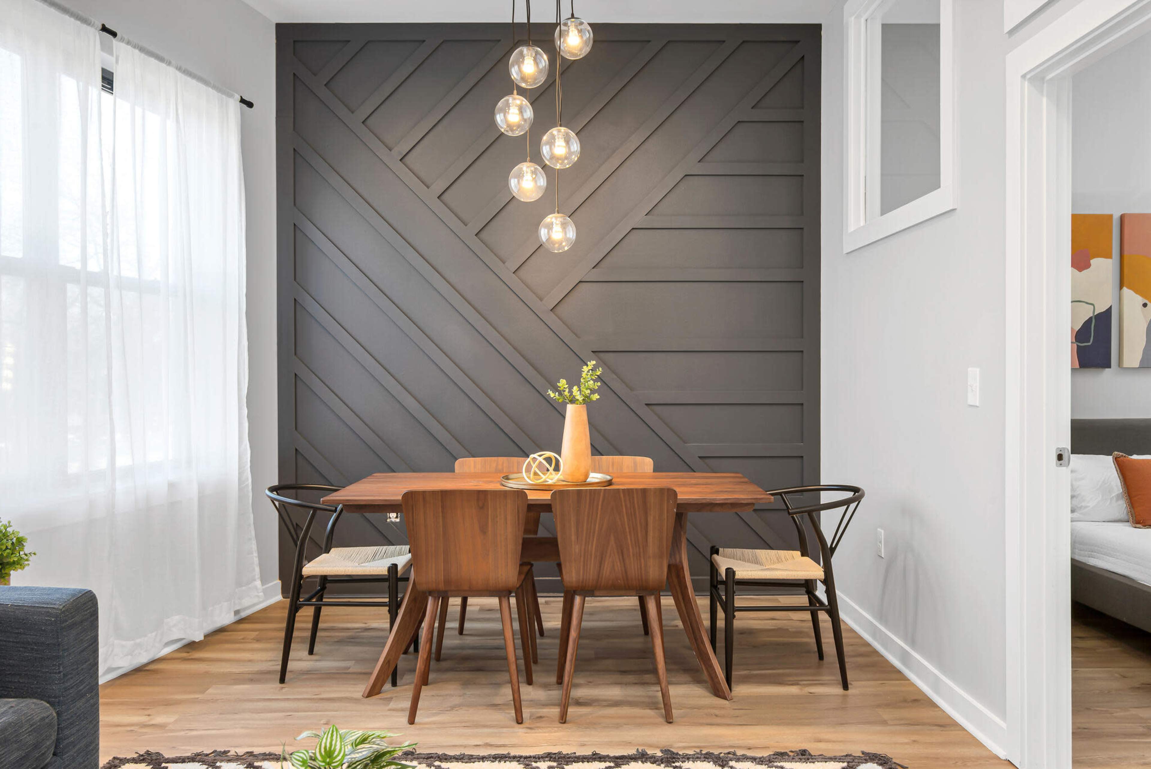 A modern dining area features a wooden table surrounded by chairs, with a accent wall displaying a geometric design and pendant lights above.
