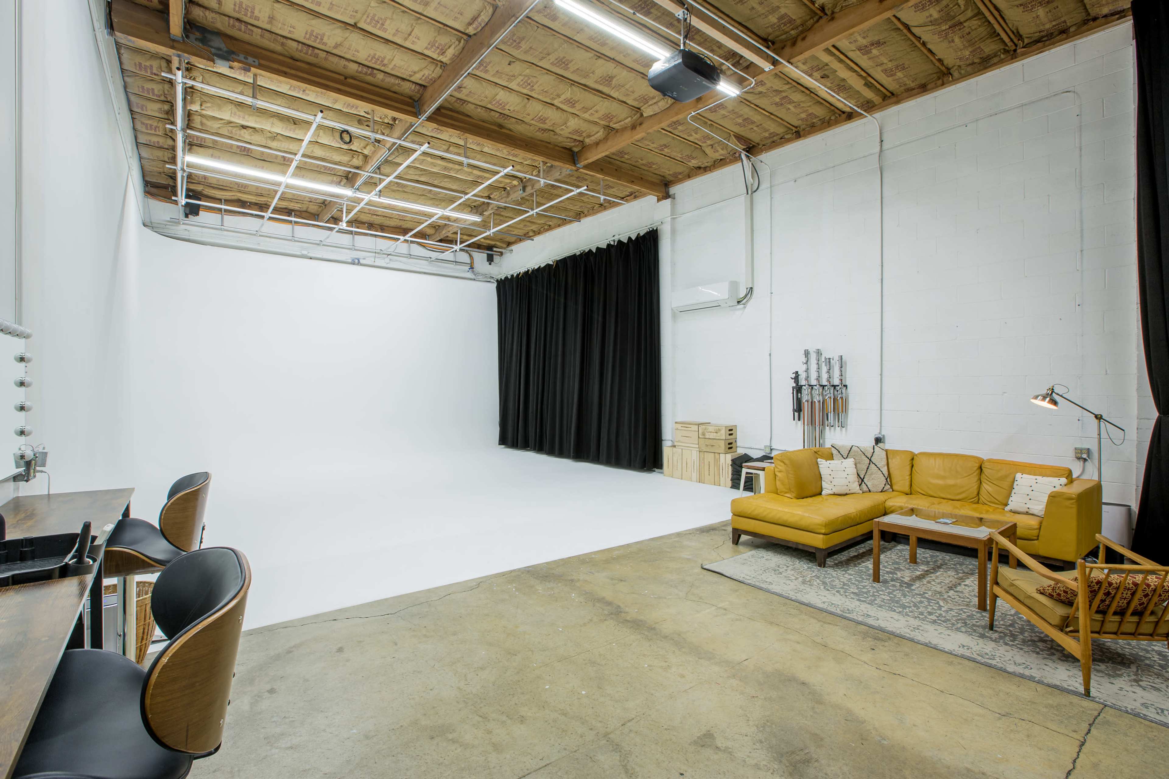 A spacious studio with a white backdrop, a black curtain on one side, a brown leather sofa, a wooden coffee table, and a small seating area near a vanity station.