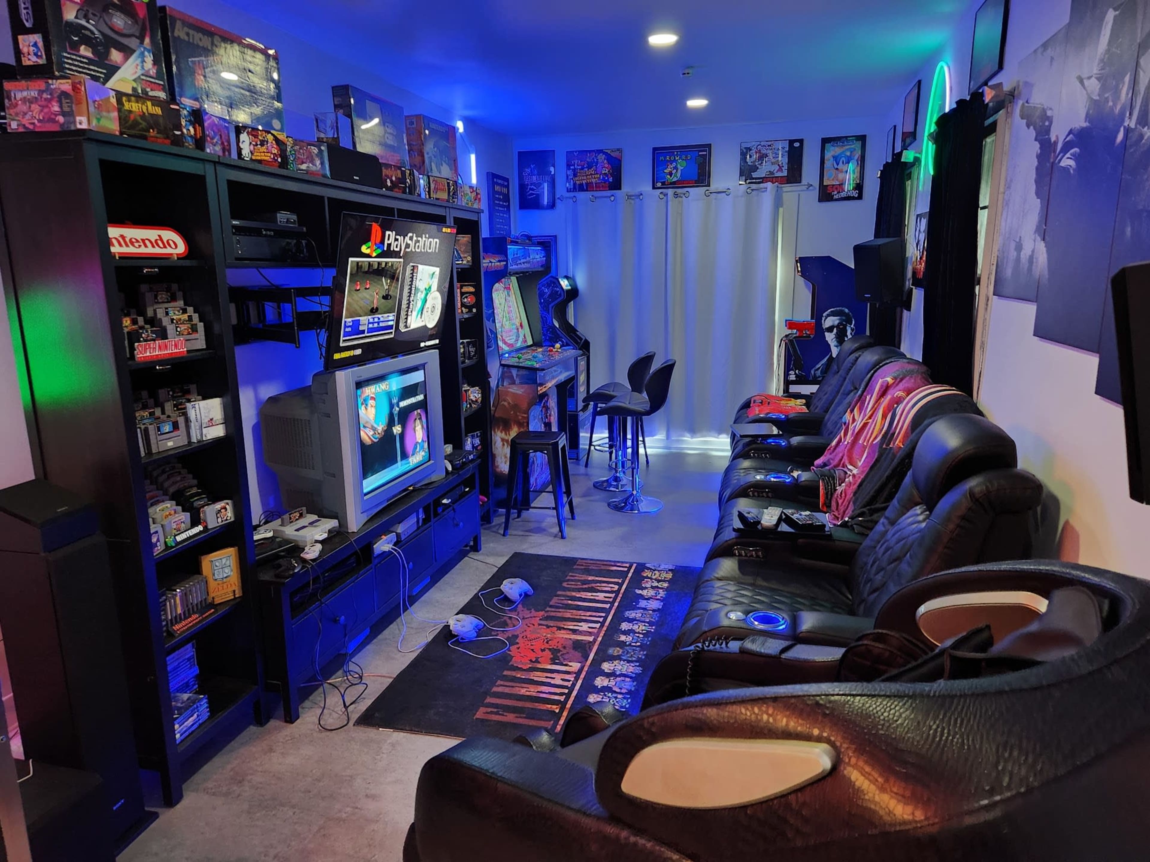 A gaming room with black leather seating, a large shelf filled with video game consoles and memorabilia, and a television displaying a game, all illuminated by colorful lights.