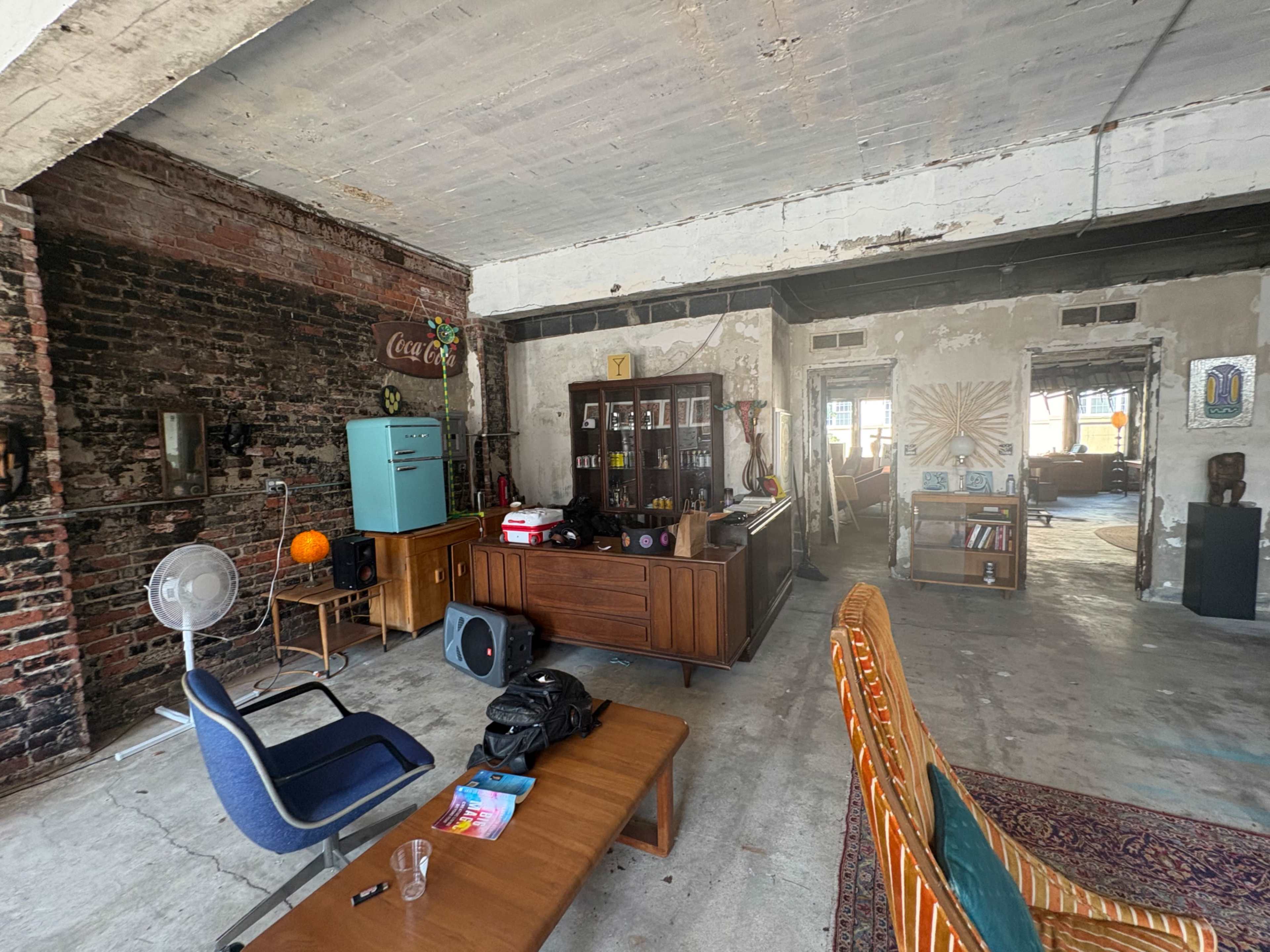 The image shows a spacious, industrial-style living room with exposed brick walls, a wooden cabinet filled with bottles, and various pieces of furniture including a couch and a table.
