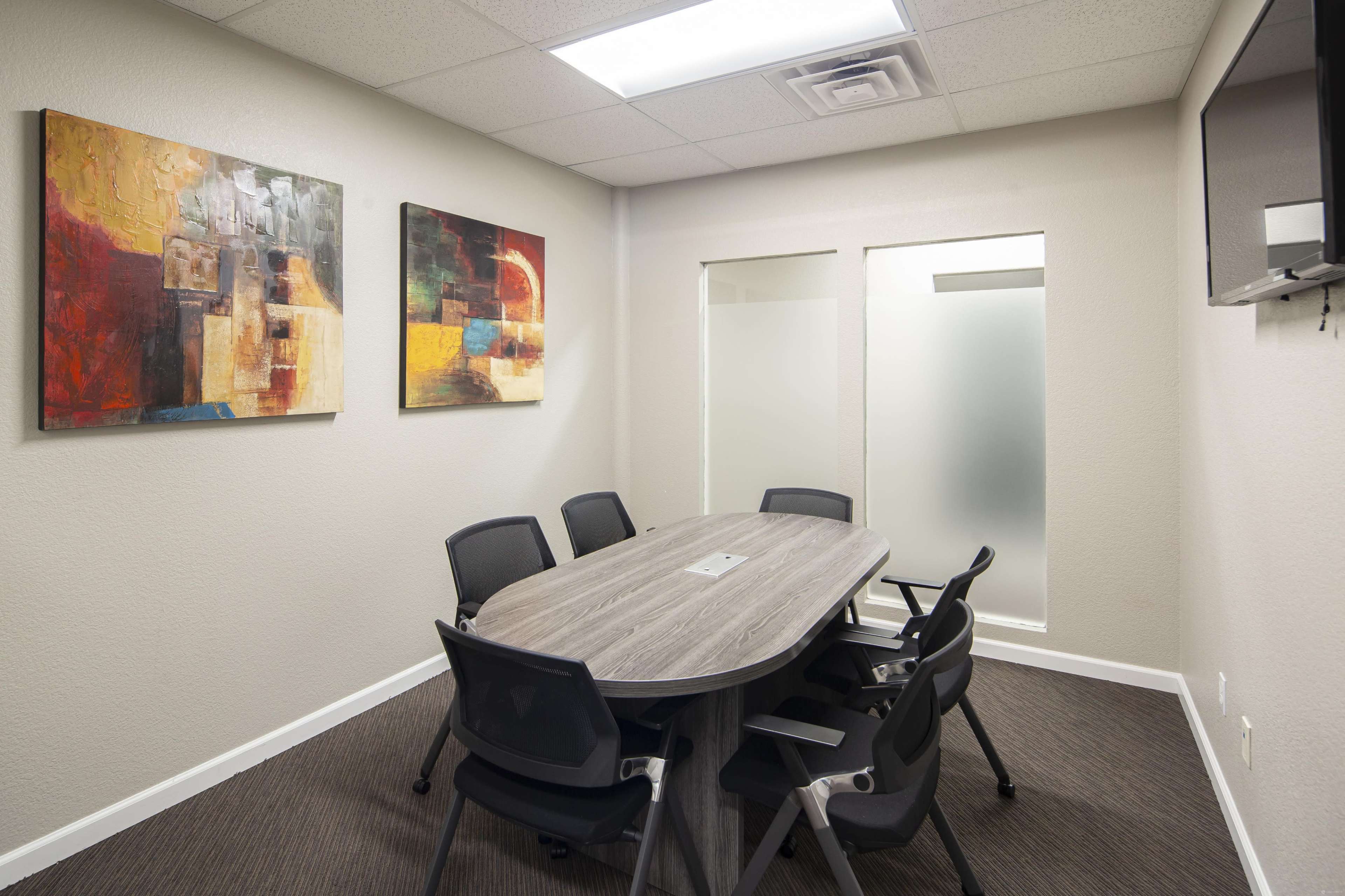 A conference room features a large oval table surrounded by black chairs, with abstract paintings on the walls and glass-paneled doors.