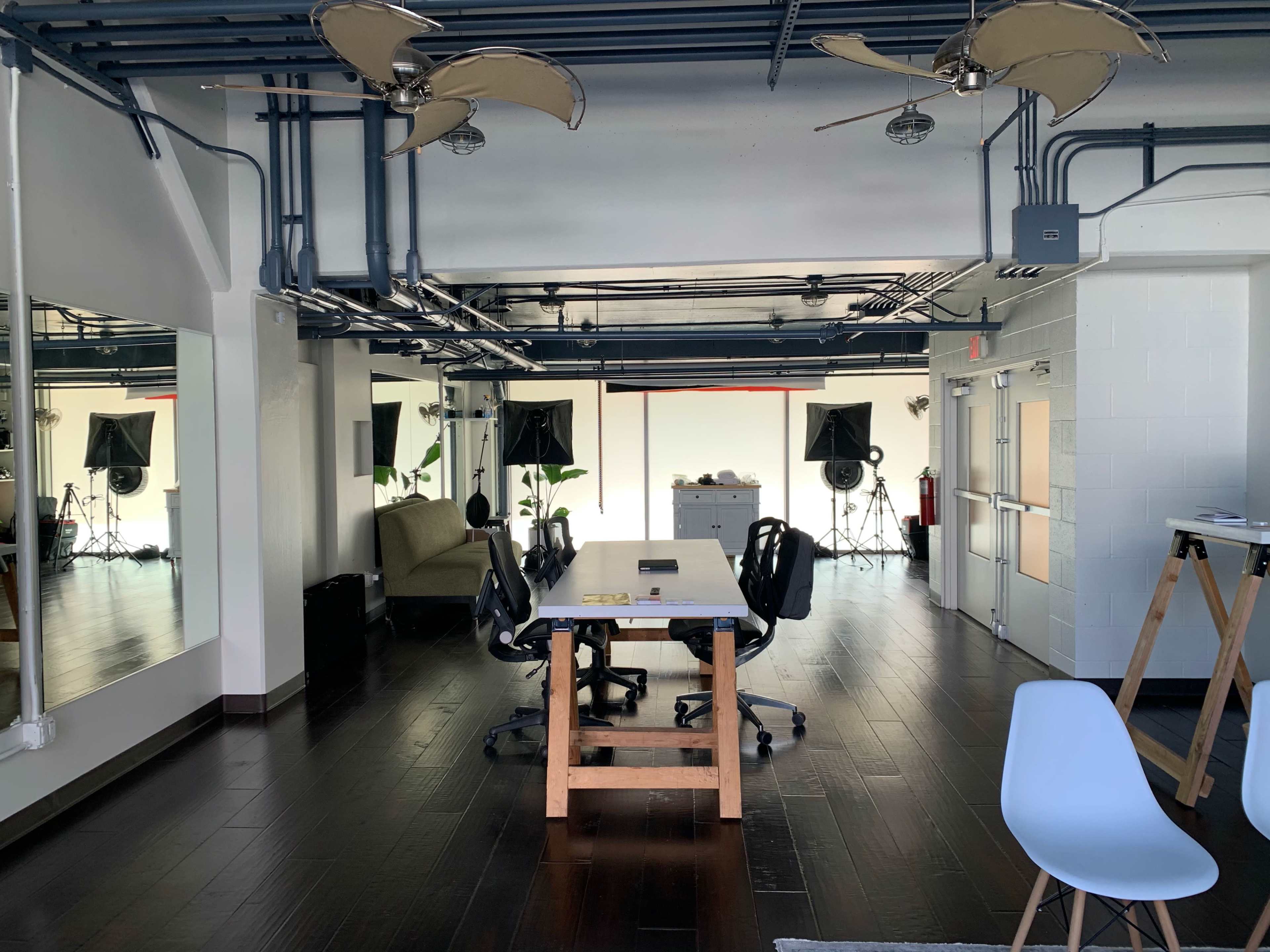 A modern office space with a large table, several chairs, two ceiling fans, and equipment set up for filming in the background.