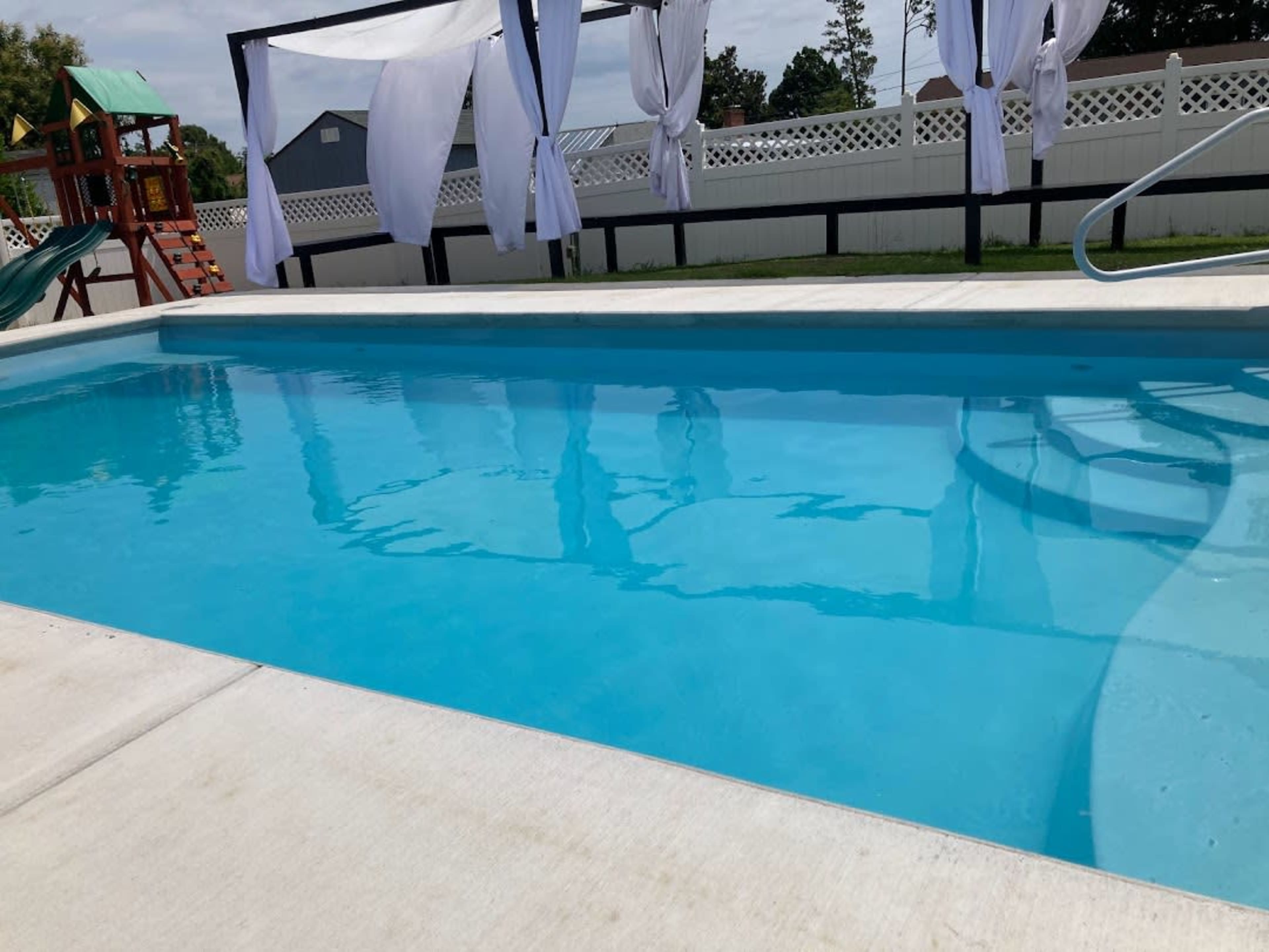 A clear swimming pool surrounded by white drapes and a play structure in the background.