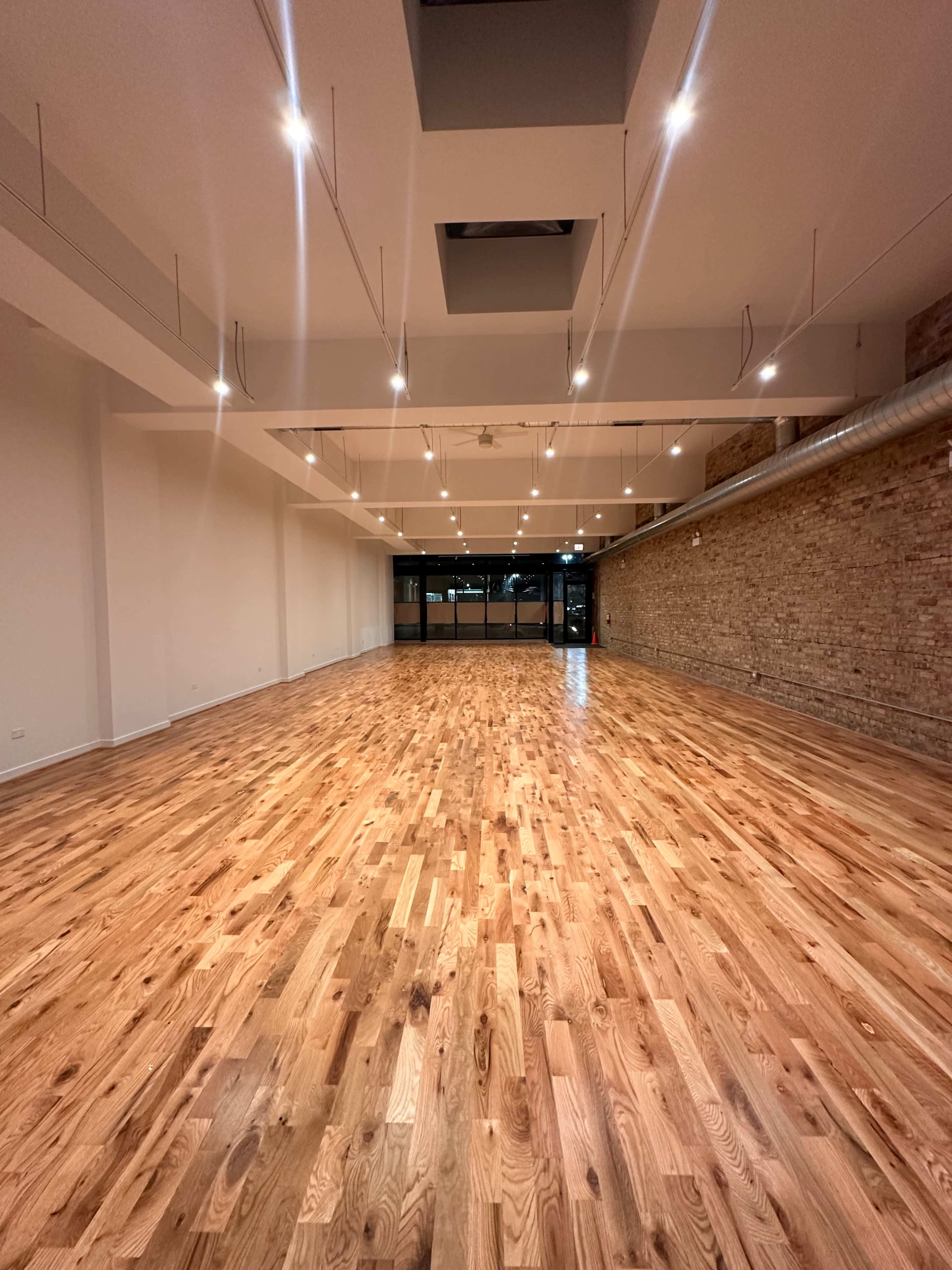 The image shows a spacious interior with polished wooden flooring, large windows allowing natural light, and modern lighting fixtures overhead.