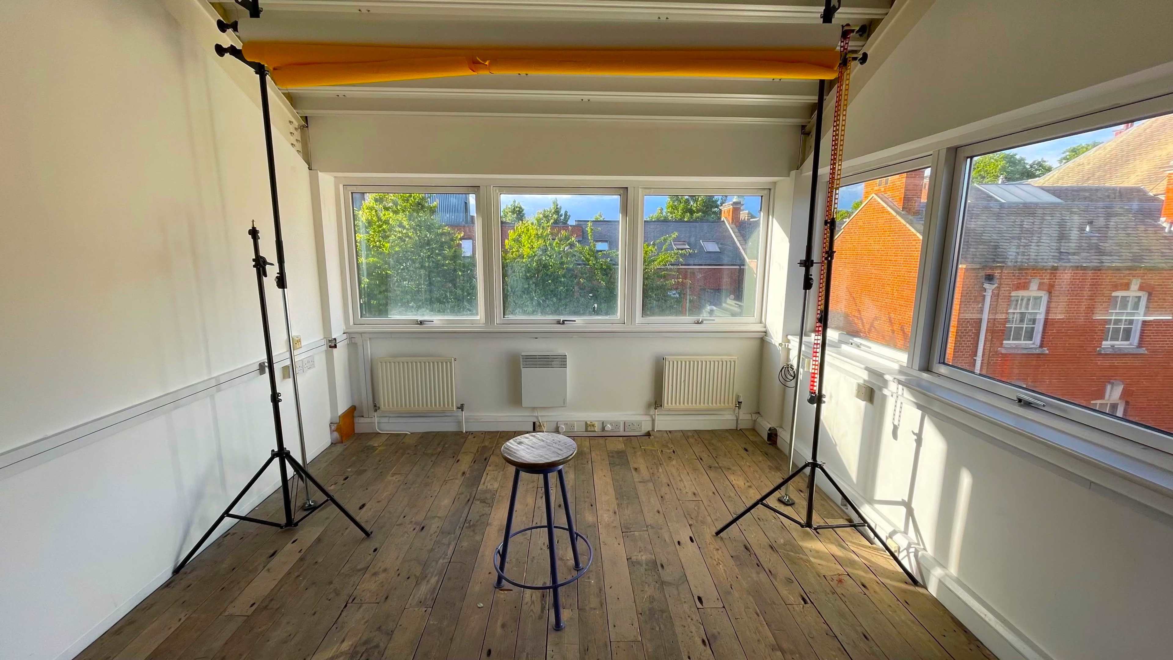 A brightly lit room with wooden flooring, featuring a stool centered under two lighting stands and large windows.
