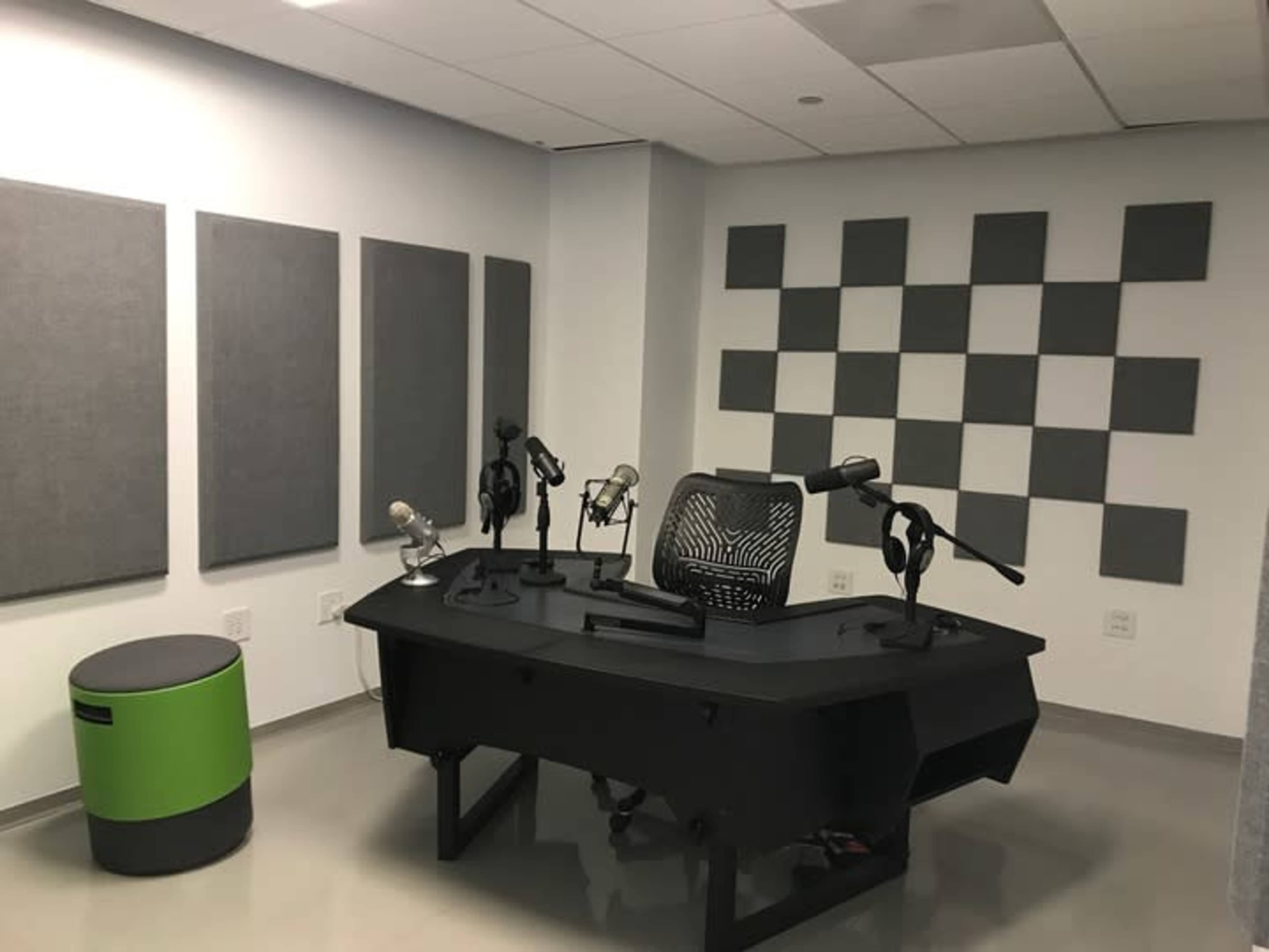 The image shows a modern podcast or recording studio featuring a black desk with multiple microphones, a swivel chair, and gray sound-absorbing panels on the walls.