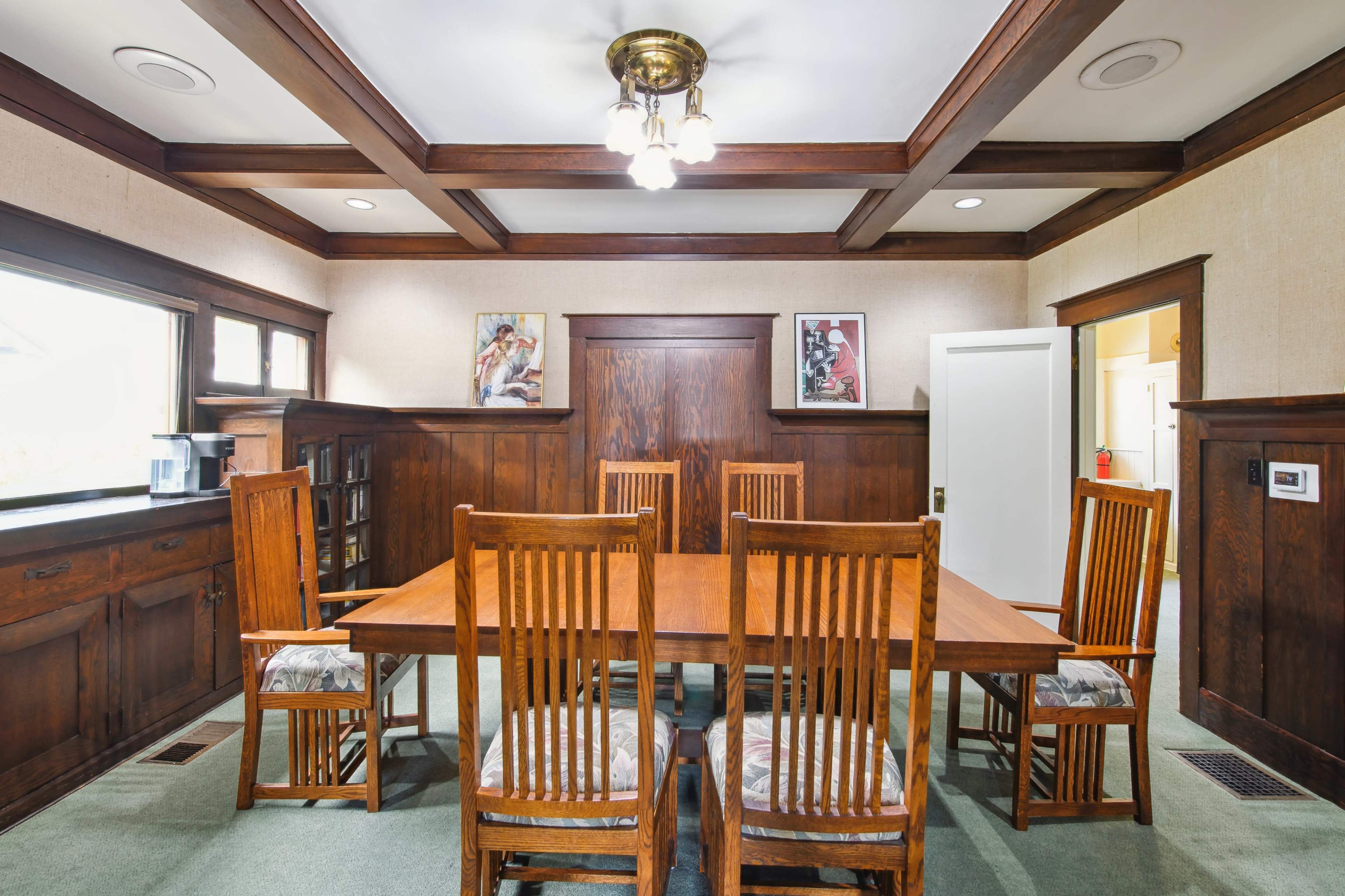A wooden dining room with a large table surrounded by six chairs, featuring exposed beam ceilings and framed artwork on the walls.