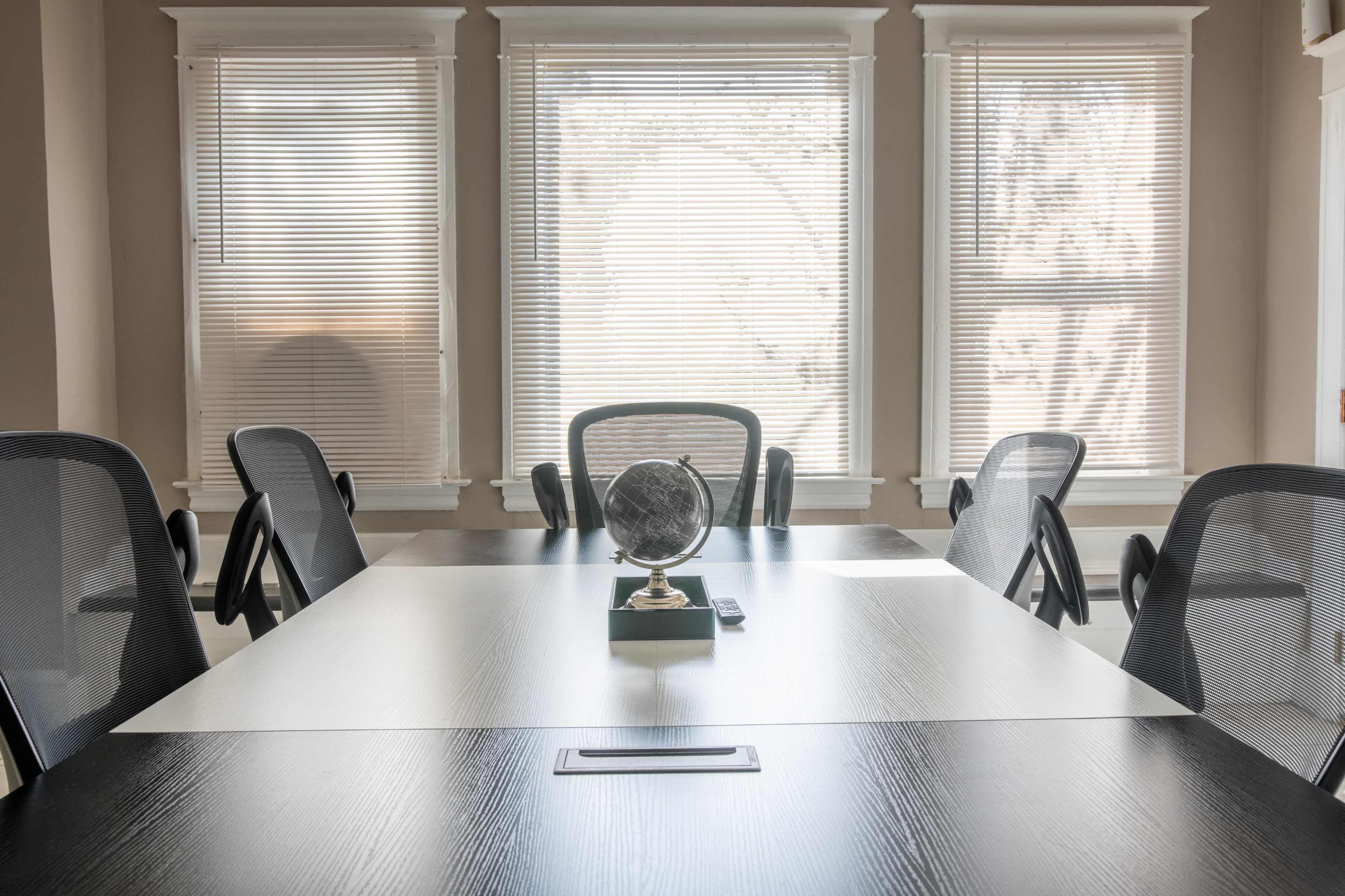 A conference room features a large table with a globe on a stand, surrounded by four black mesh chairs and bright windows with blinds.