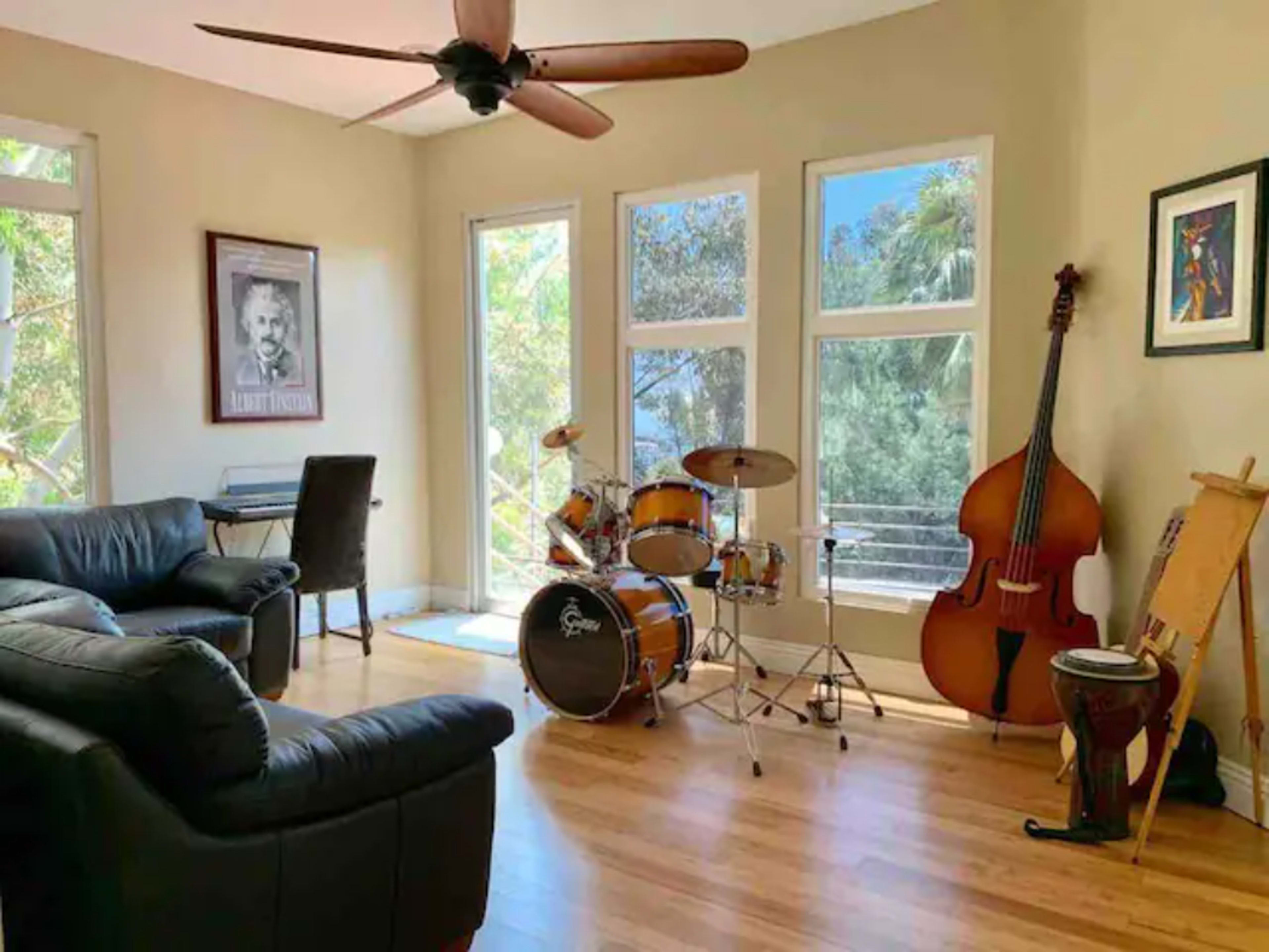 A musical room featuring a black leather couch, a drum set, a double bass, and an easel, with large windows providing a view of greenery outside.