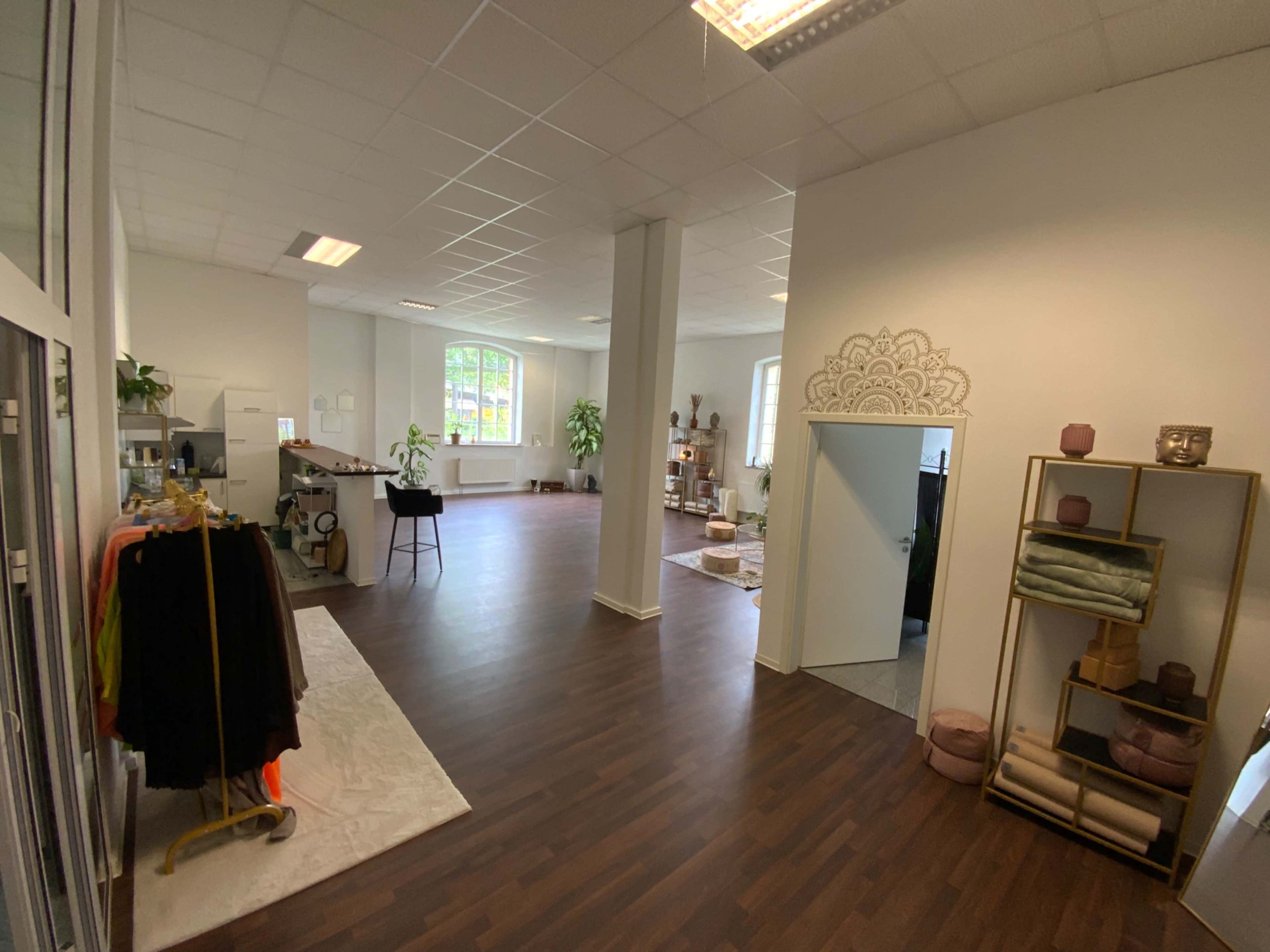 The image shows a spacious, well-lit interior of a retail or studio space with plants and decorative elements, featuring a clothing rack on the left and a shelving unit on the right.