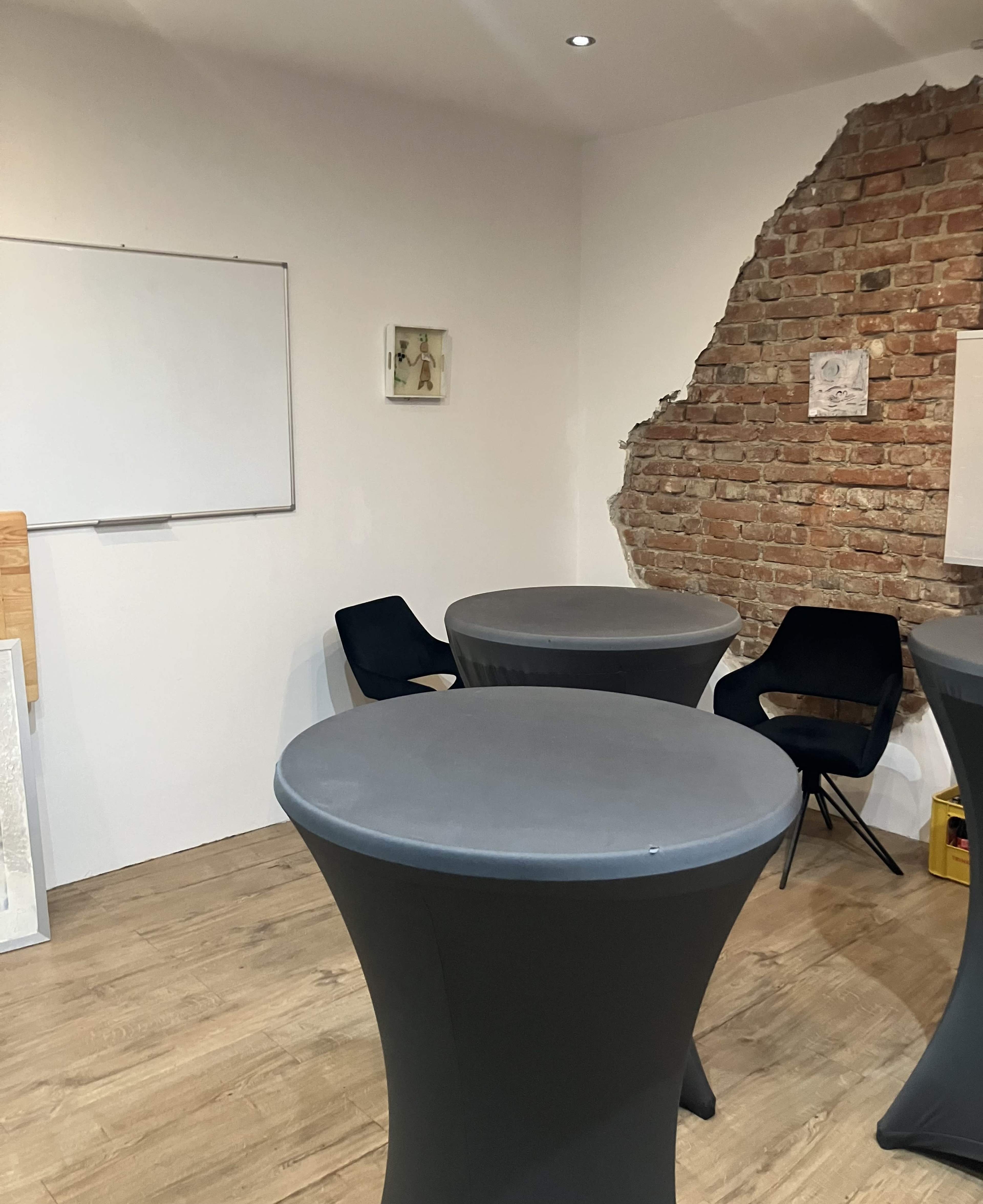 The image shows a small room with three gray round tables and two black chairs, featuring a brick wall on one side and a whiteboard on another.