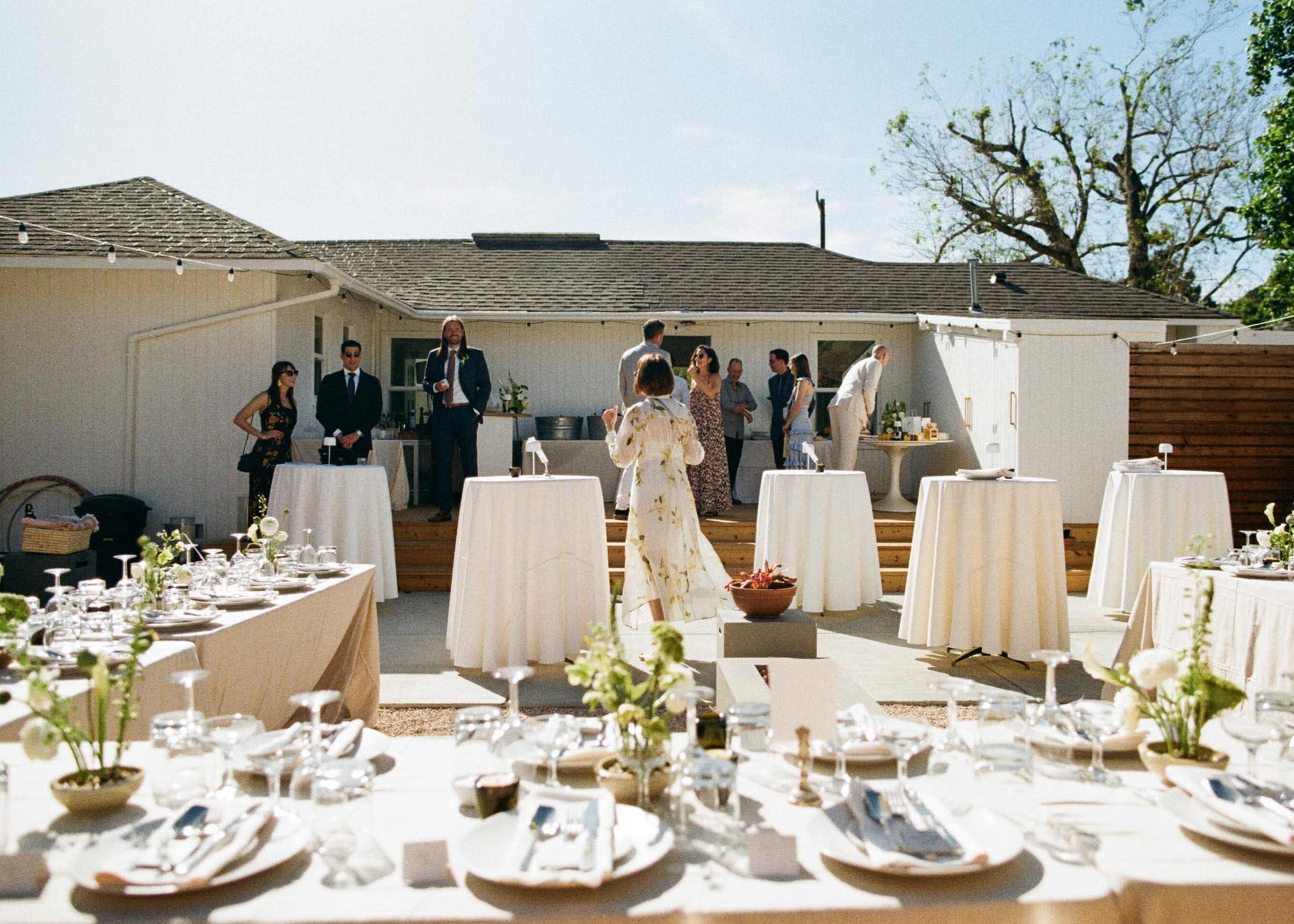 An outdoor gathering is set with several dining tables, surrounded by guests and a house in the background.