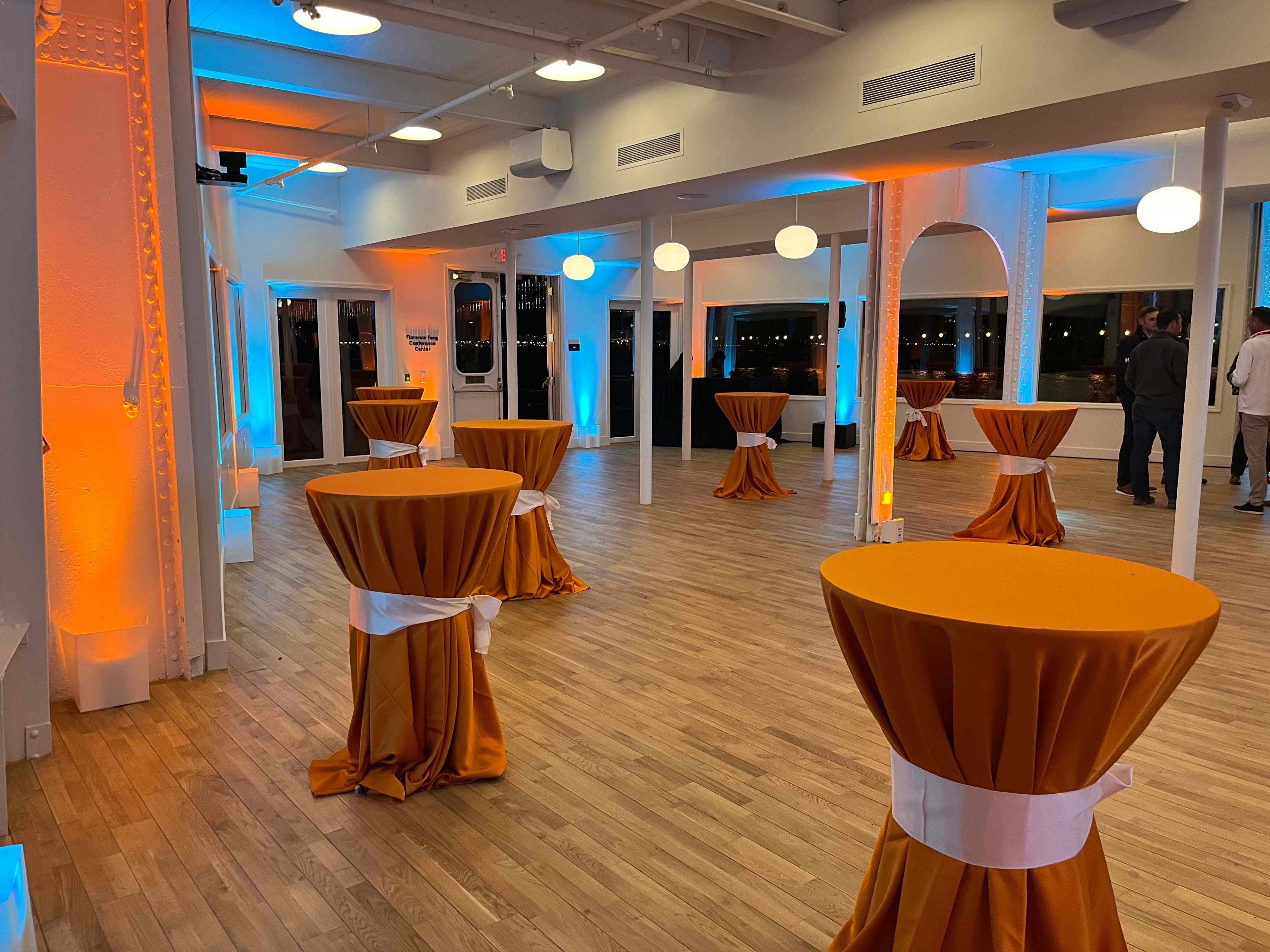 The image shows a spacious event room with tall, round tables covered in orange tablecloths, illuminated by soft blue and orange lighting.