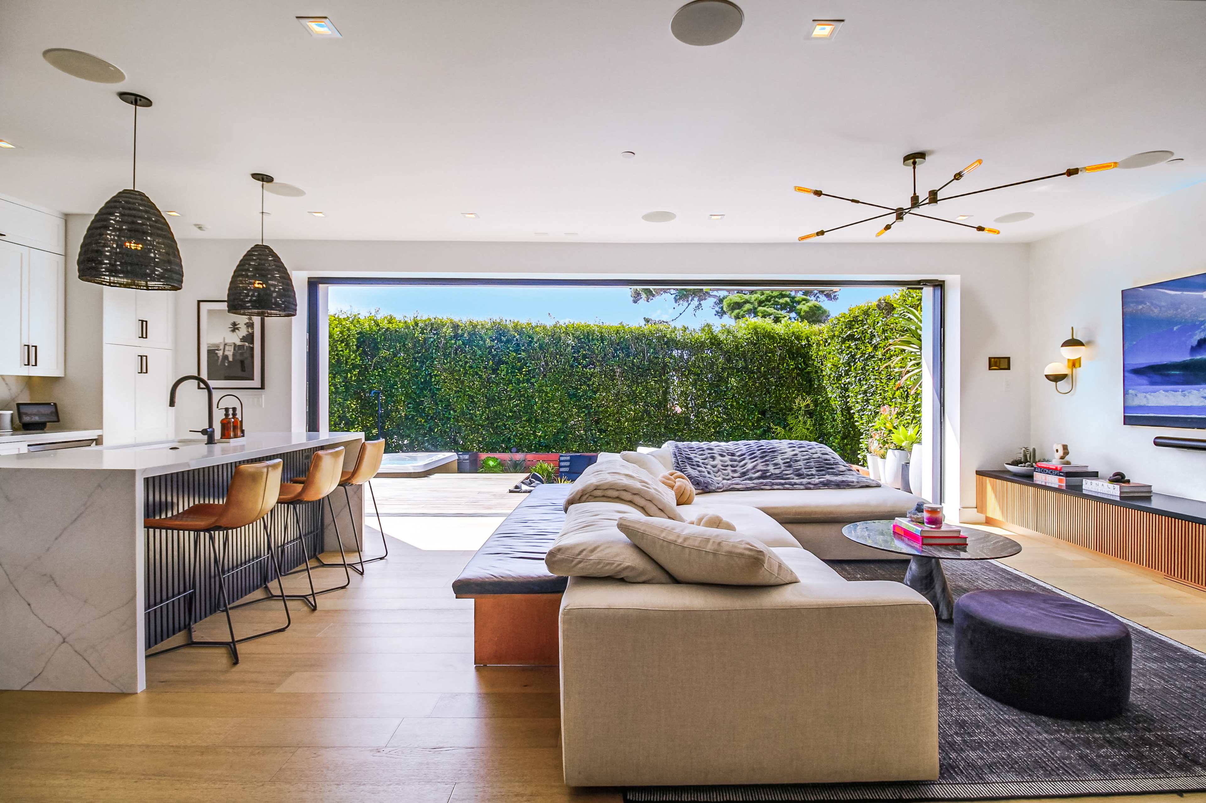 A modern open-concept living space features a large sectional sofa, a minimalist kitchen with bar seating, and expansive glass doors leading to a patio with greenery.