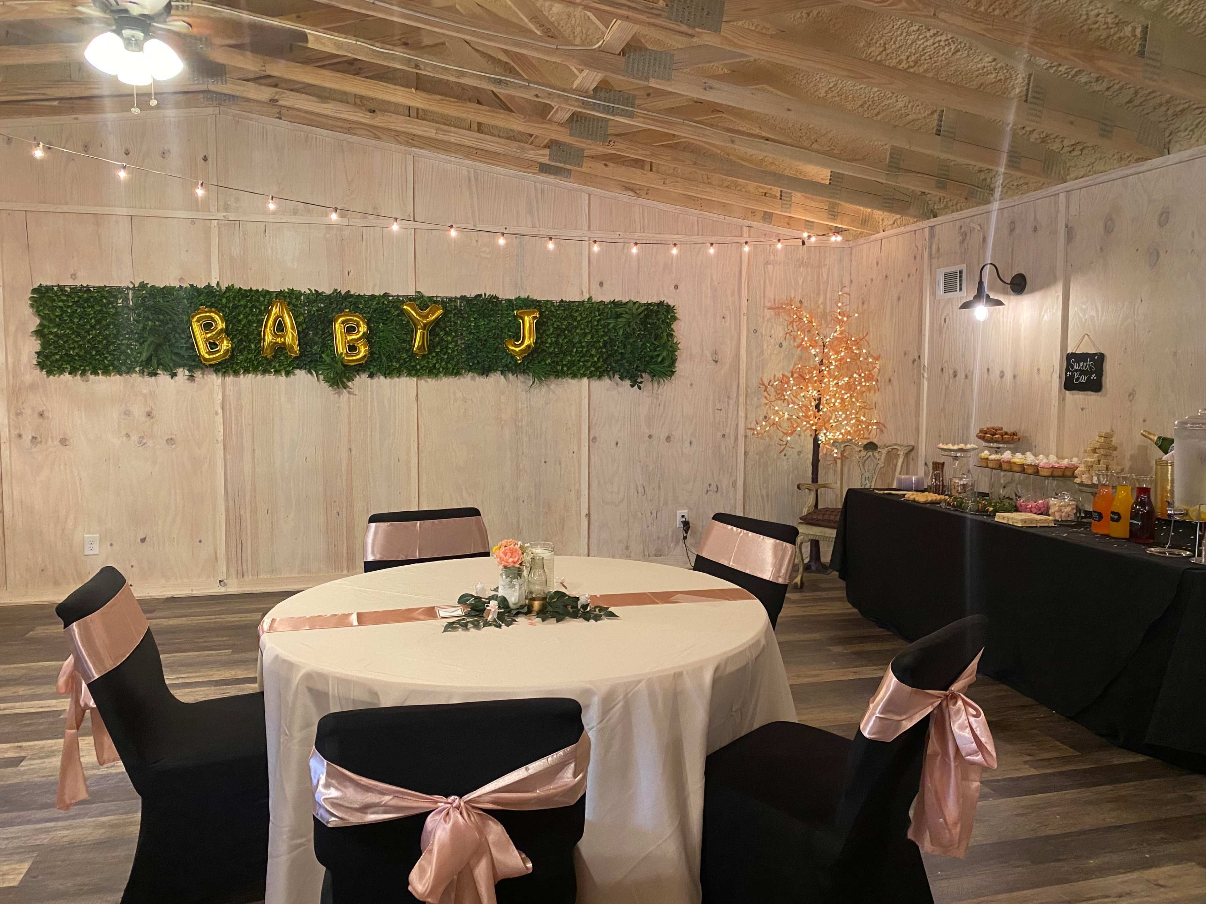 An indoor event space decorated for a celebration, featuring a round table with a white tablecloth and pink accents, a green wall with the letters "BABY J," and a buffet table with various treats.