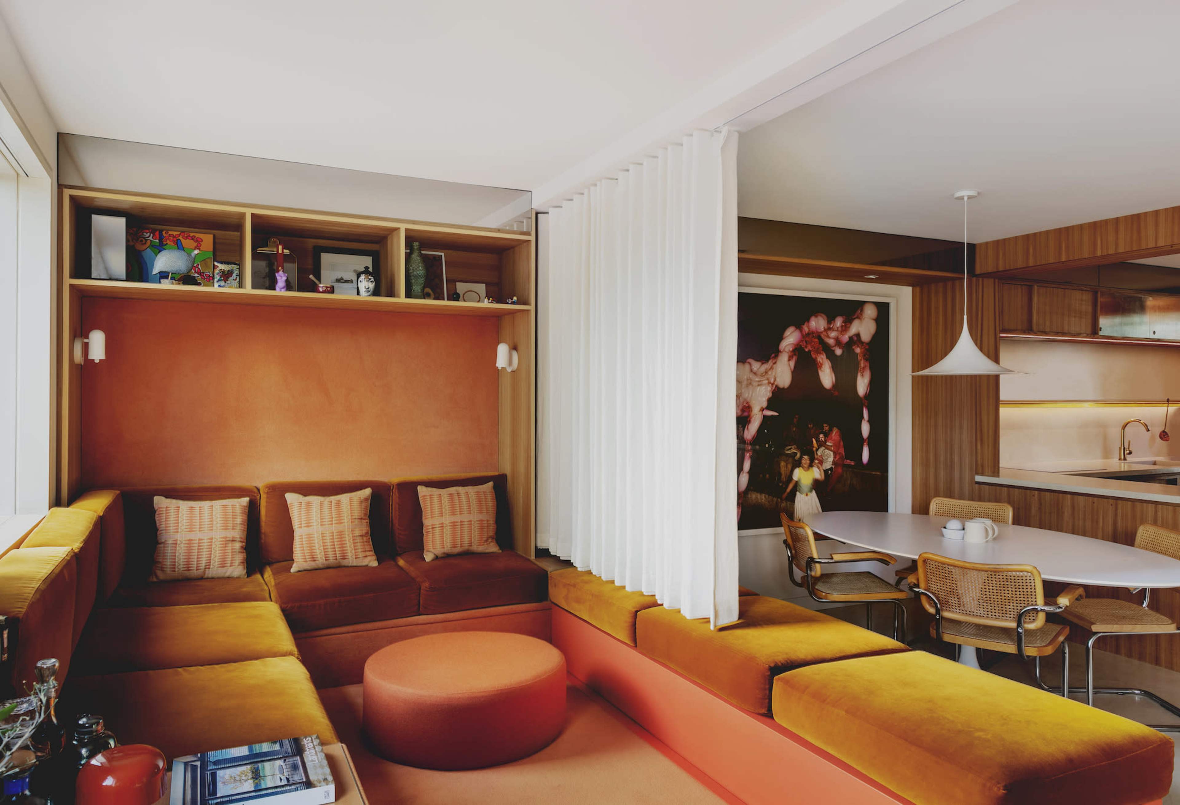 The image depicts a modern living room with an orange and yellow sectional sofa, a round ottoman, and a dining area featuring a round table and woven chairs, all against a backdrop of wood paneling and decorative shelves.