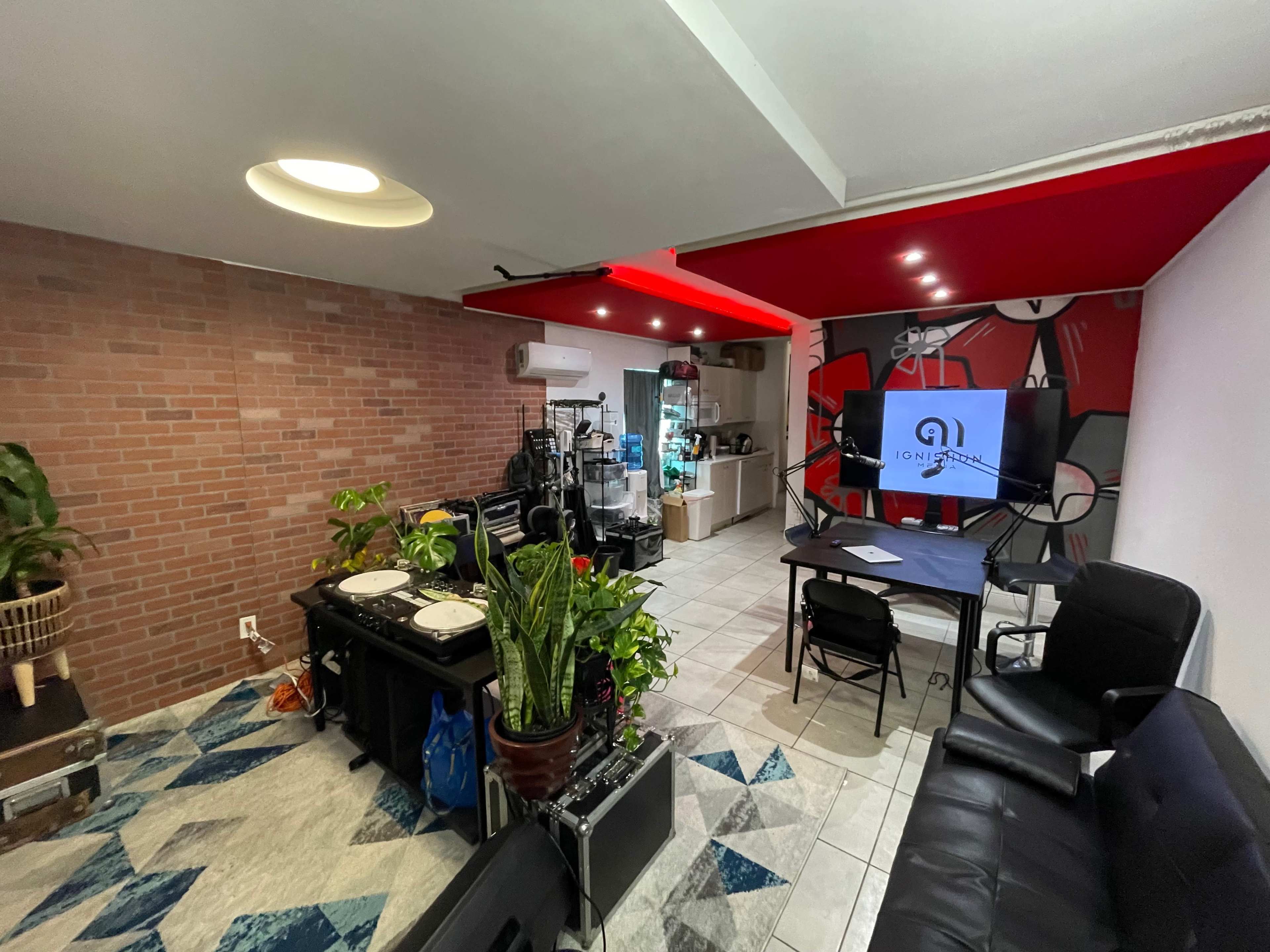 A multi-purpose room featuring a kitchenette, a lounge area with a black sofa, a table, and equipment for music or video production.