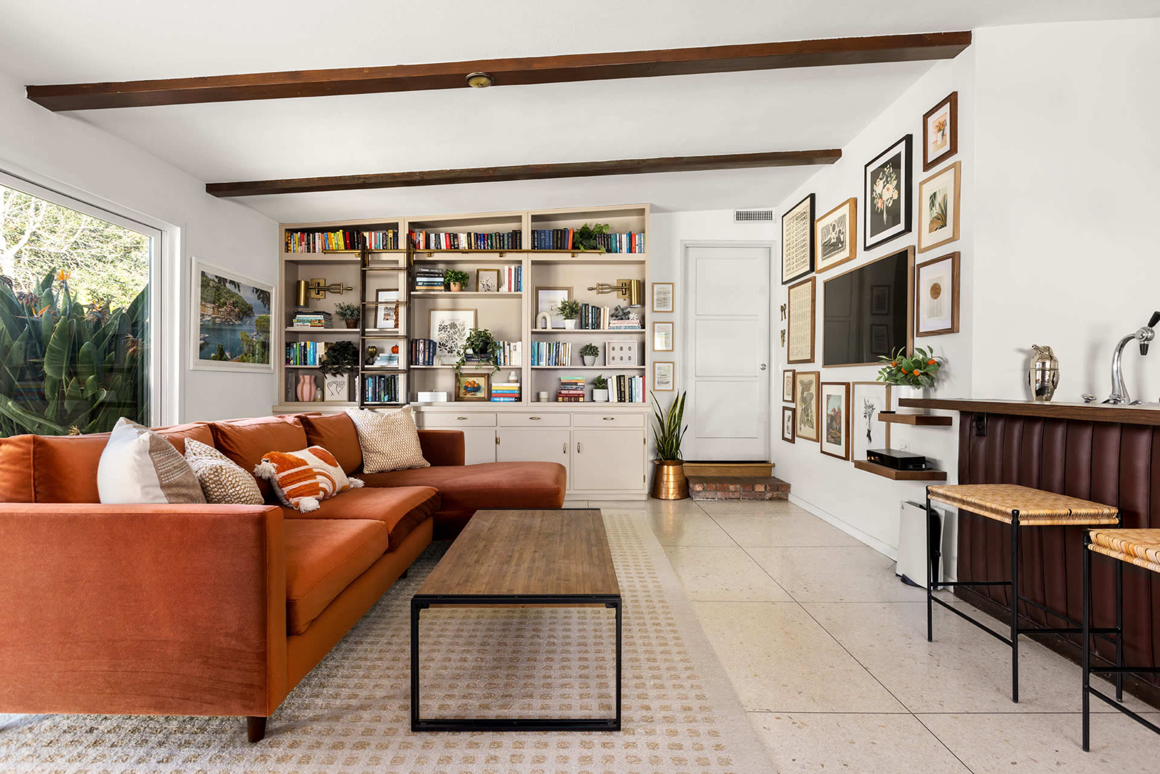 A cozy living room with an orange sofa, a wooden coffee table, a bookshelf filled with books, and a small bar area against a wall adorned with framed pictures.