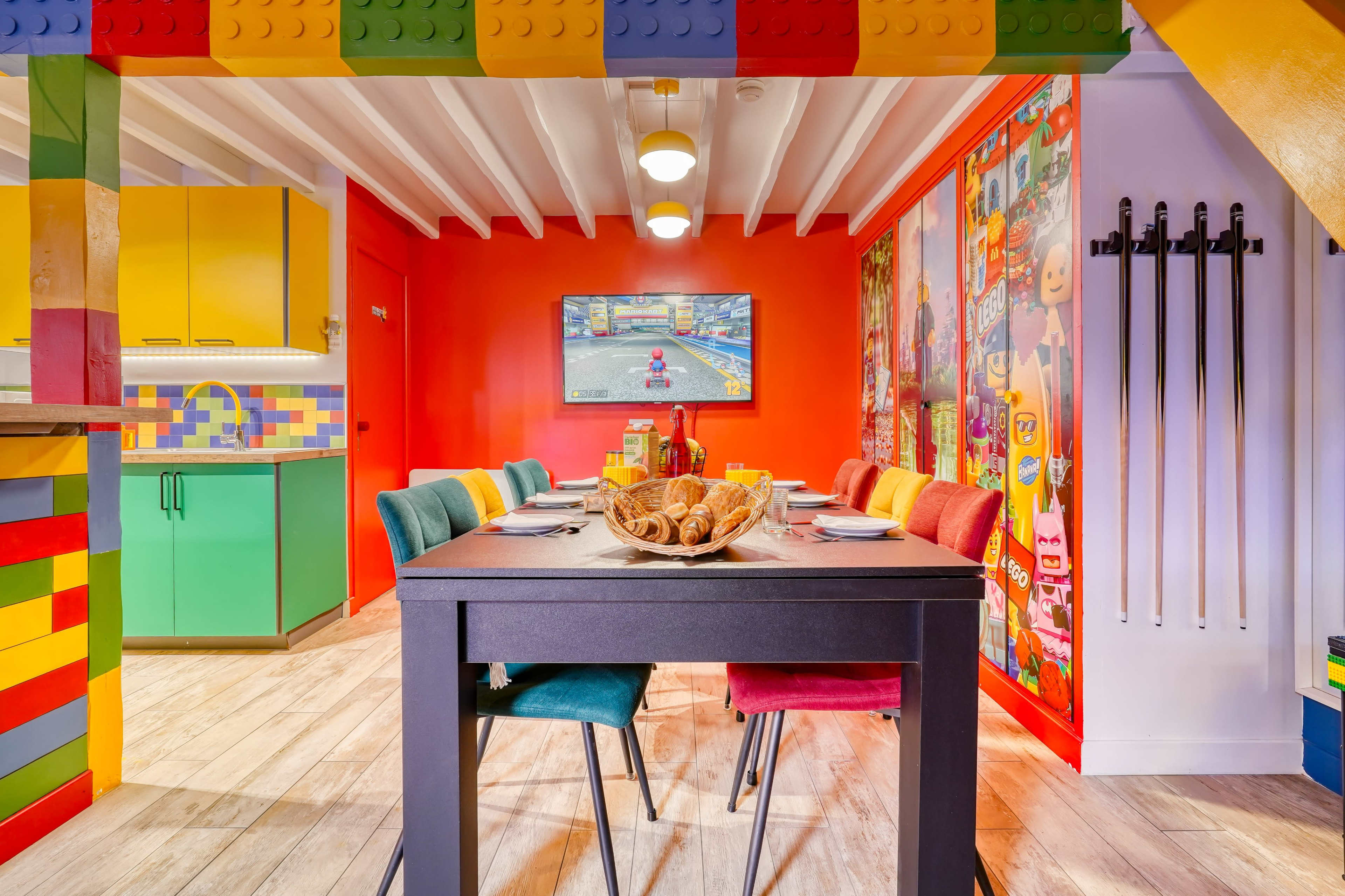The image shows a colorful dining area with a table set for a meal, surrounded by chairs in various hues, and walls decorated with Lego-inspired designs and vibrant artwork.