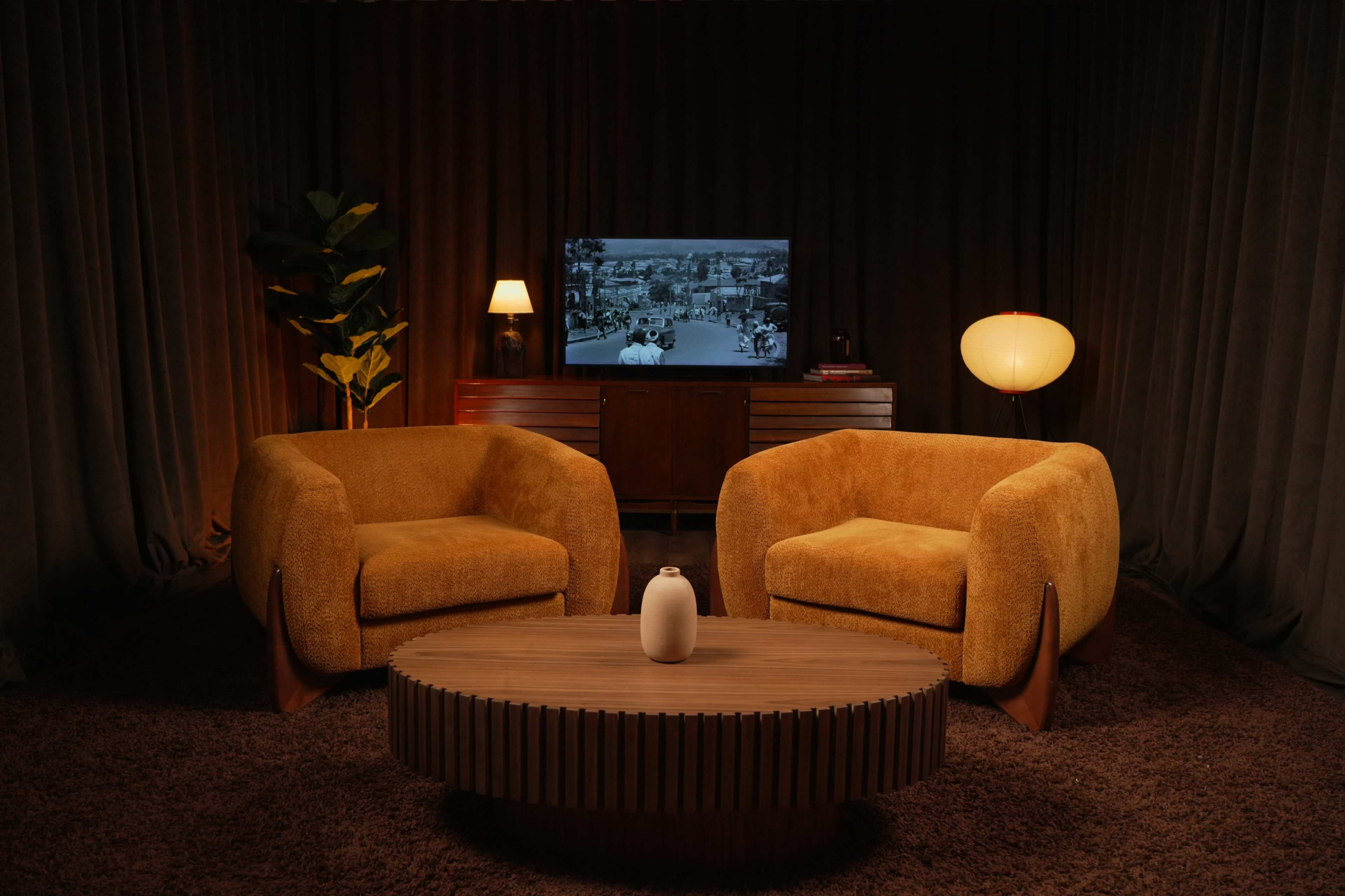 The scene features two yellow upholstered chairs facing a wooden coffee table in a dimly lit room with a television in the background.