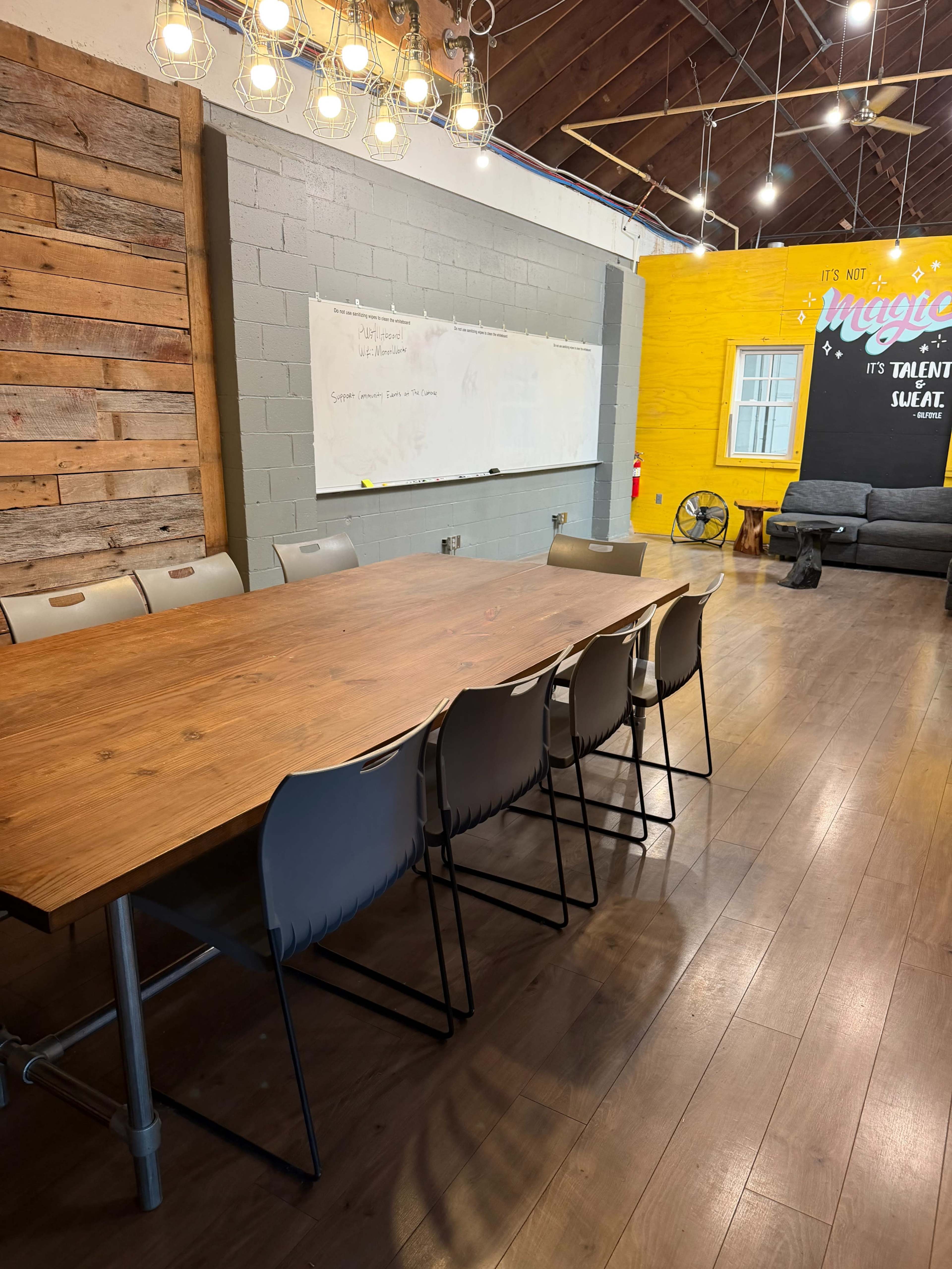 The image shows a meeting room featuring a large wooden table surrounded by several chairs, a whiteboard on the wall, and a cozy seating area with a couch.