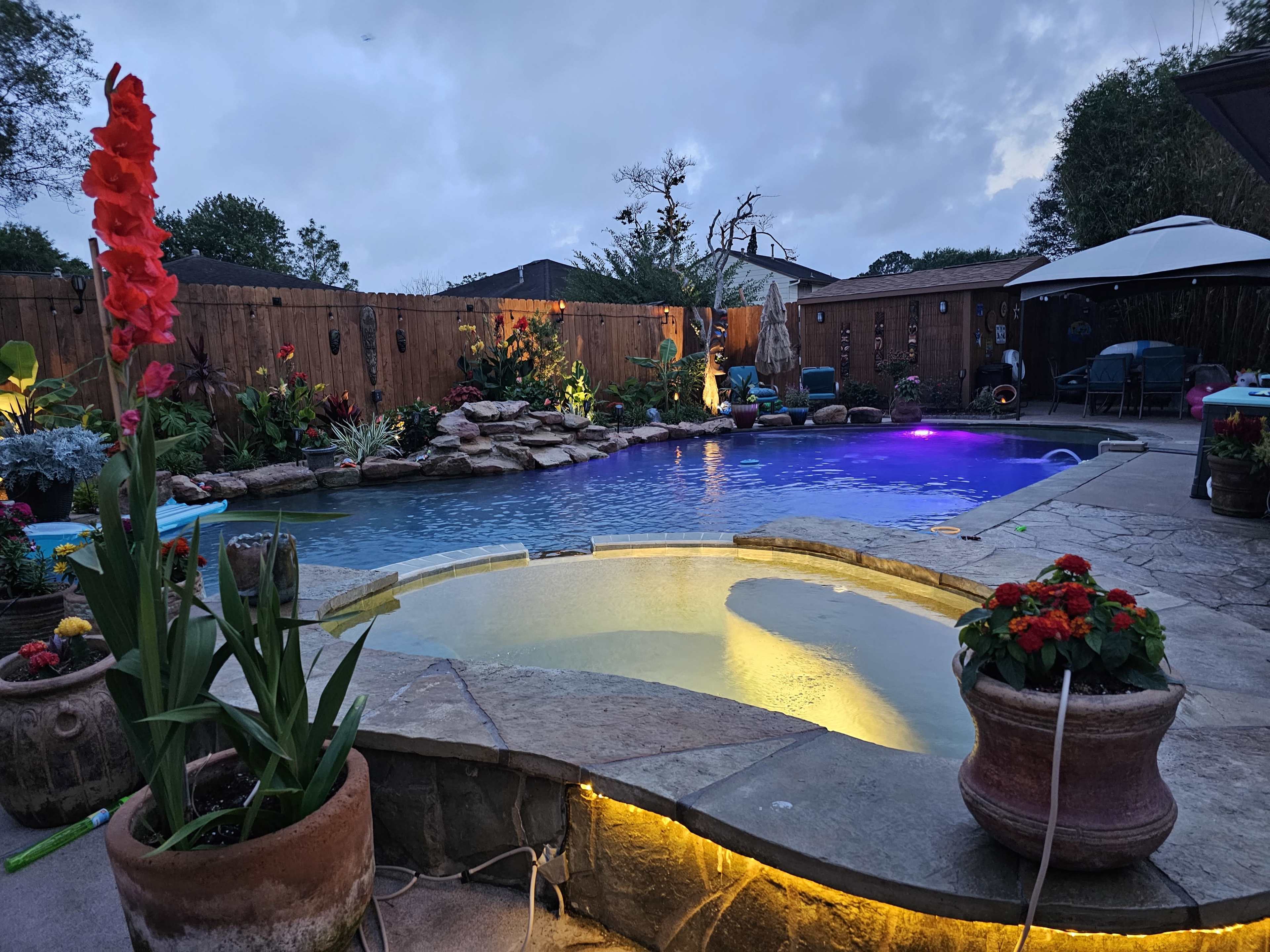 A swimming pool surrounded by landscaped gardens, with brightly colored flowers and ambient lighting under a cloudy sky.