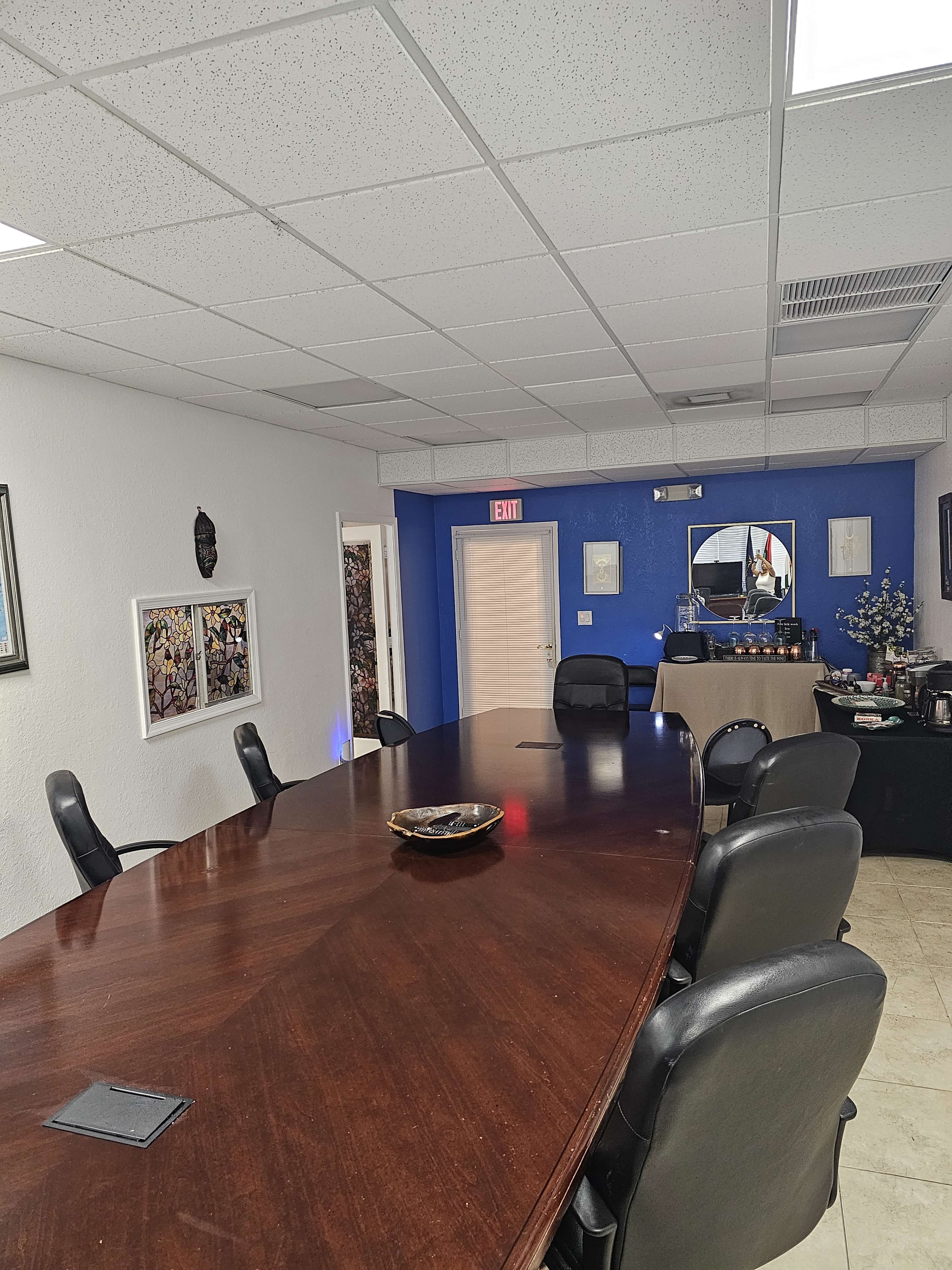 A conference room with a long wooden table surrounded by black chairs, and a blue wall adorned with framed art and a mirror.