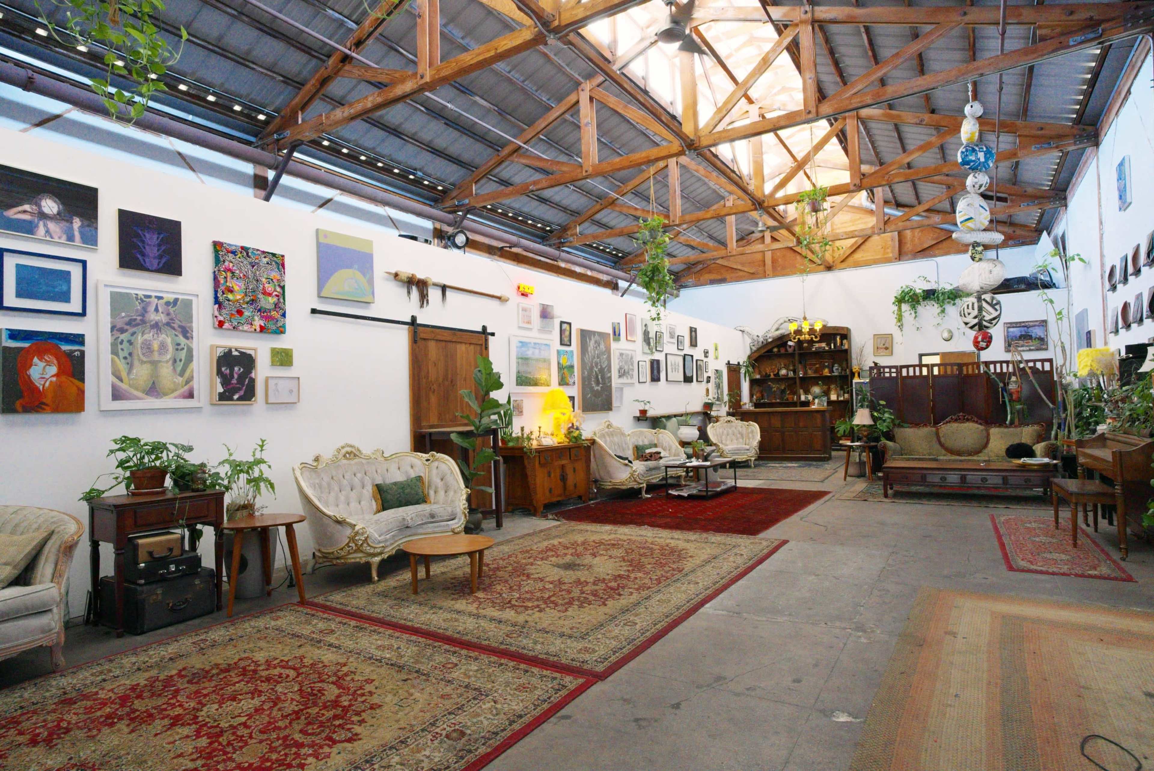 The image shows a spacious, well-lit room with wooden beams, decorated with various artworks on the walls and furnished with vintage seating and rugs.