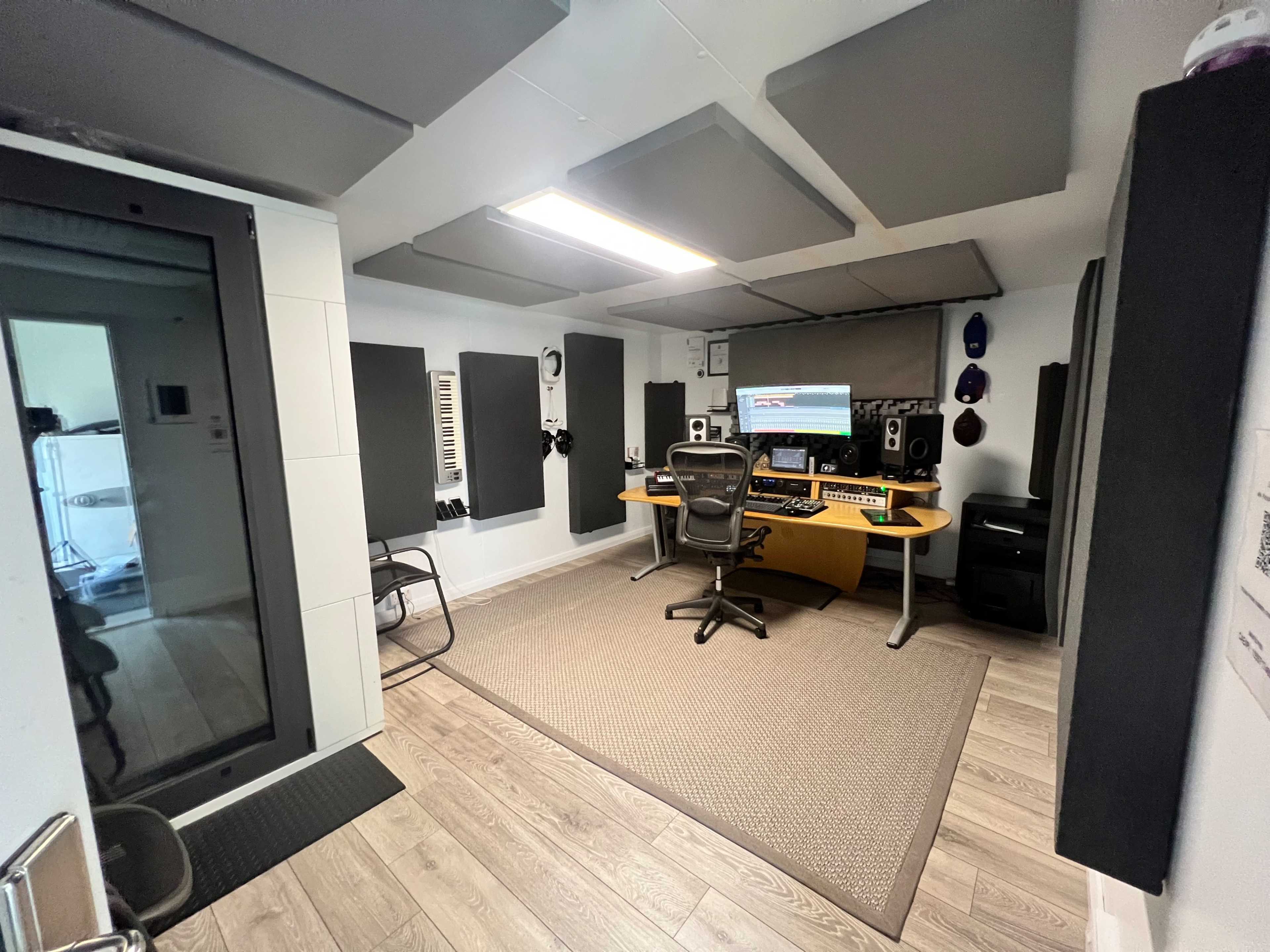 A modern music studio featuring a computer workstation, soundproof panels on the walls, and a glass isolation booth.