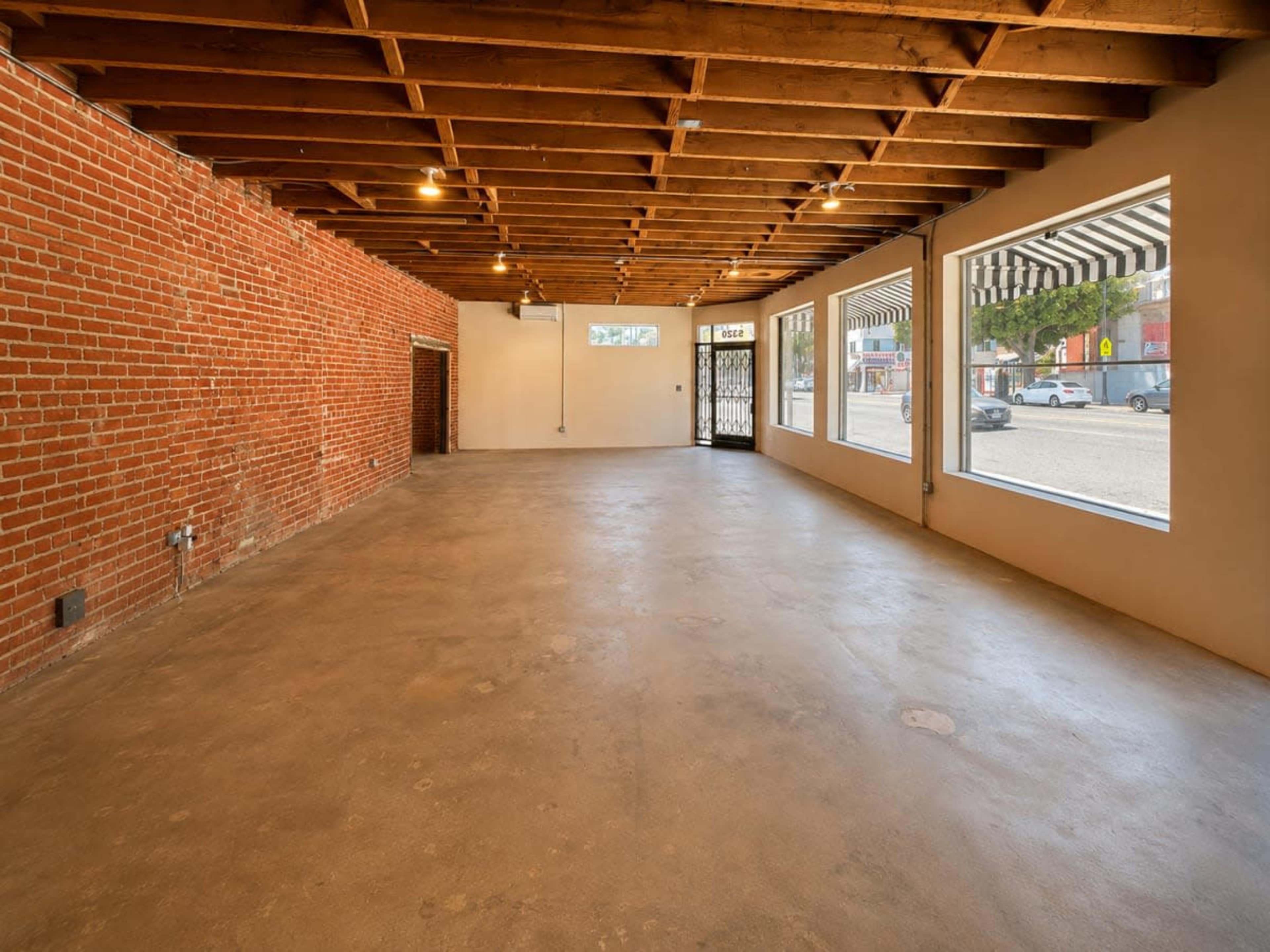 The image shows an empty interior space with brick walls, wooden beams, and large windows that allow natural light to enter.