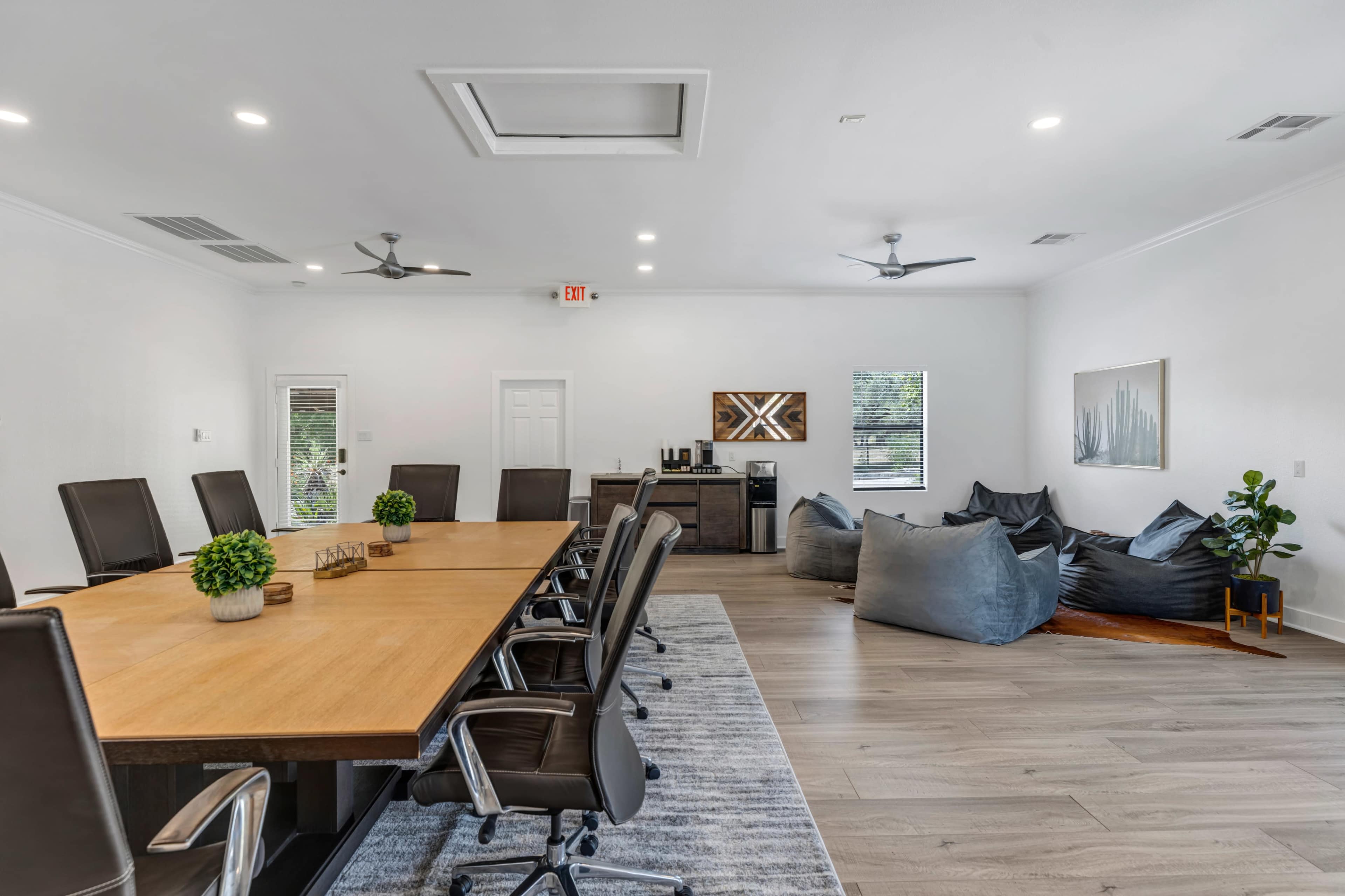 A spacious conference room features a long wooden table surrounded by black office chairs, with bean bag seating and a coffee area against the walls.