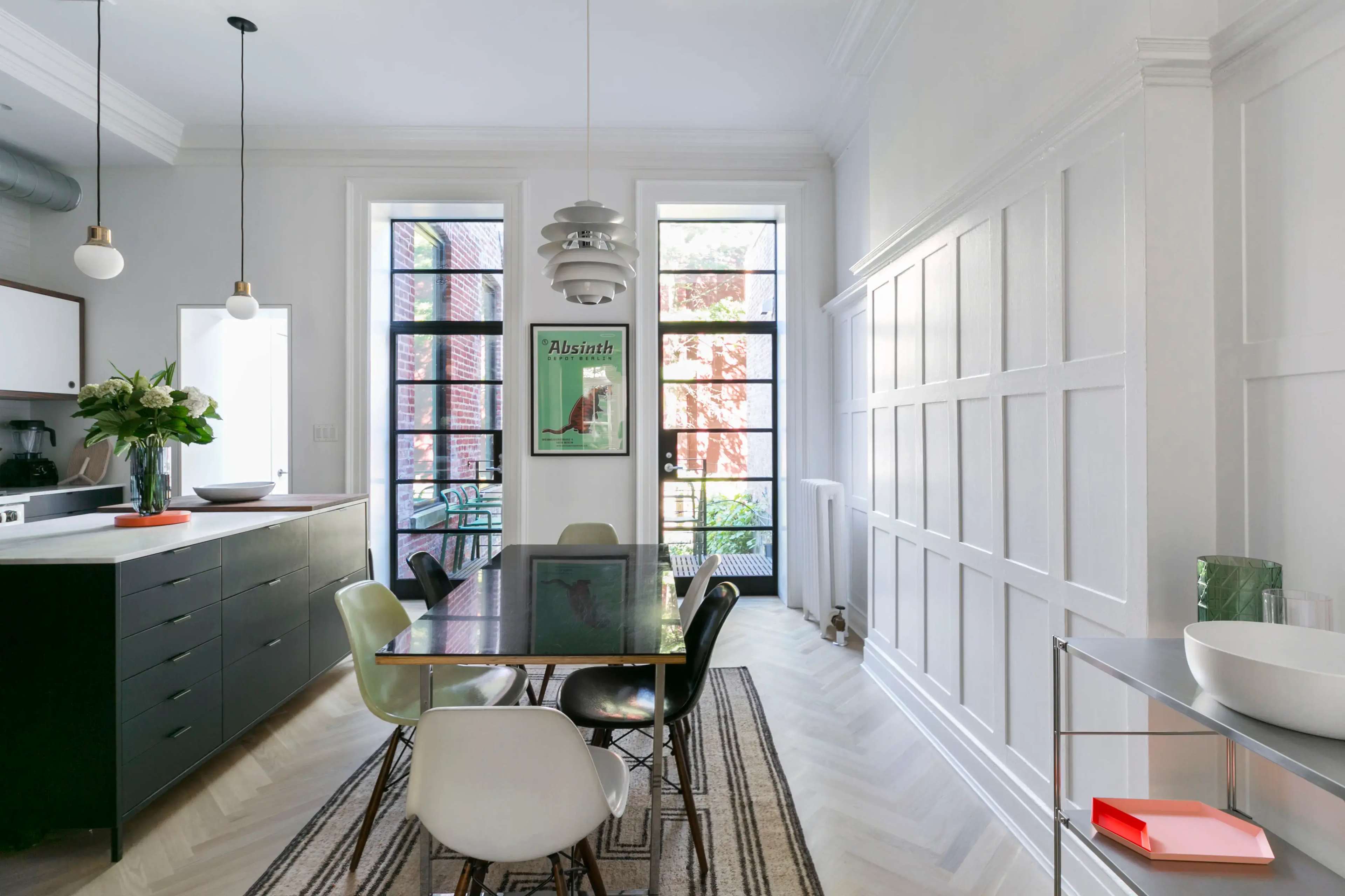 A modern dining area with a glass table surrounded by various colored chairs, large windows letting in natural light, and a minimalist kitchen in the background.